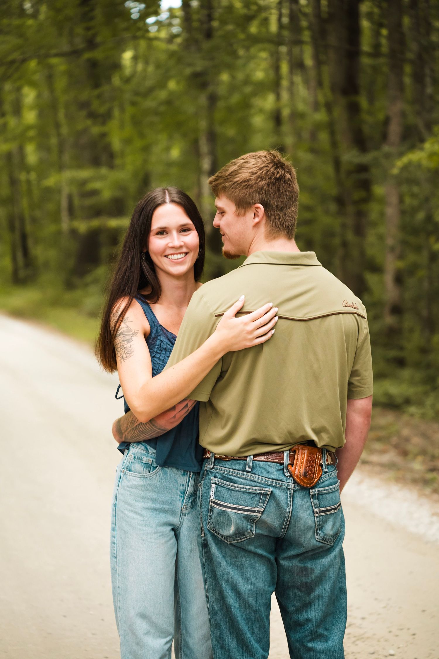 A smiling brunette woman embraces a man in a green polo shirt on a tree-lined gravel road.