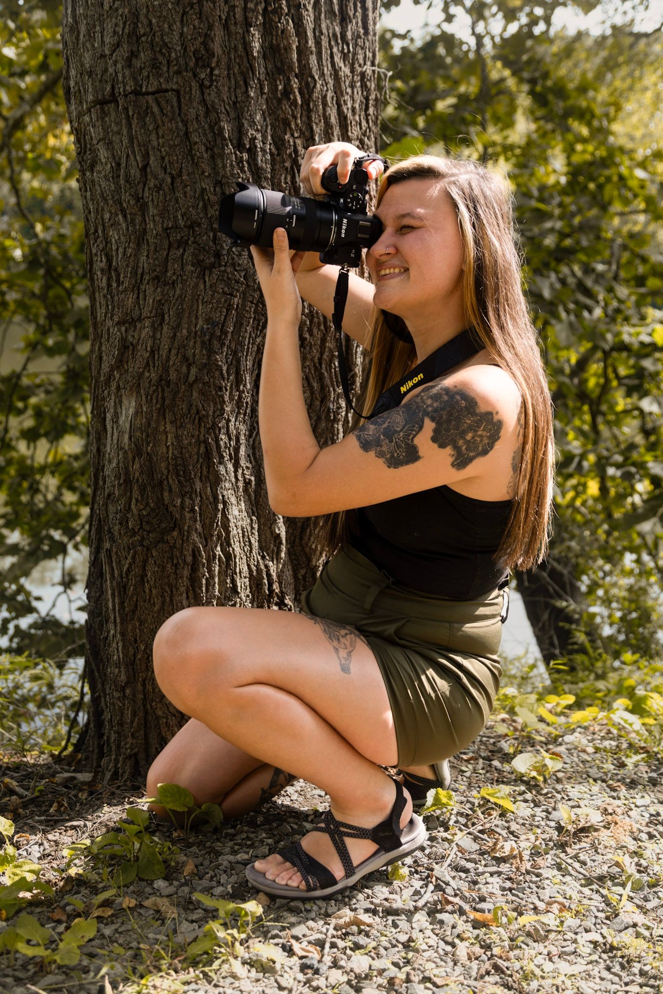 Smiling woman with tattoos crouching by a tree, photographing nature with a DSLR camera outdoors.