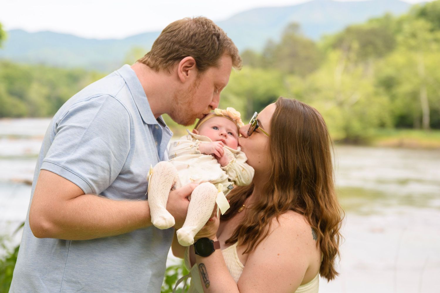 Parents kissing their newborn baby outdoors near a river with green mountains in the background.