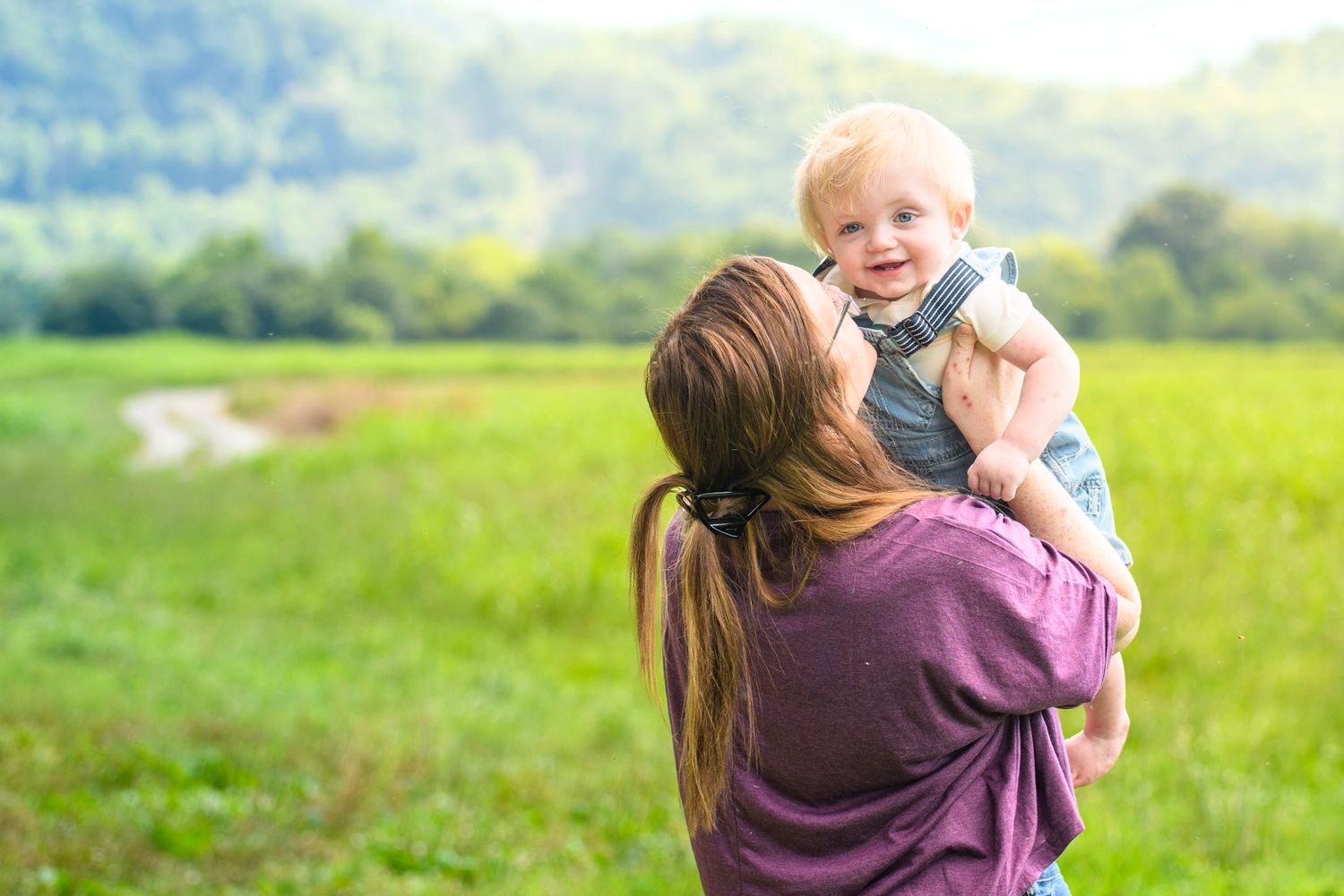 A mother lifts her smiling toddler boy up in the air in a lush green field on a sunny day.