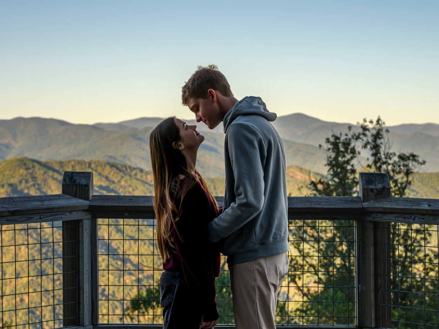 A couple embraces on a wooden overlook, sharing an intimate moment with layered mountain ridges in the background at sunset.