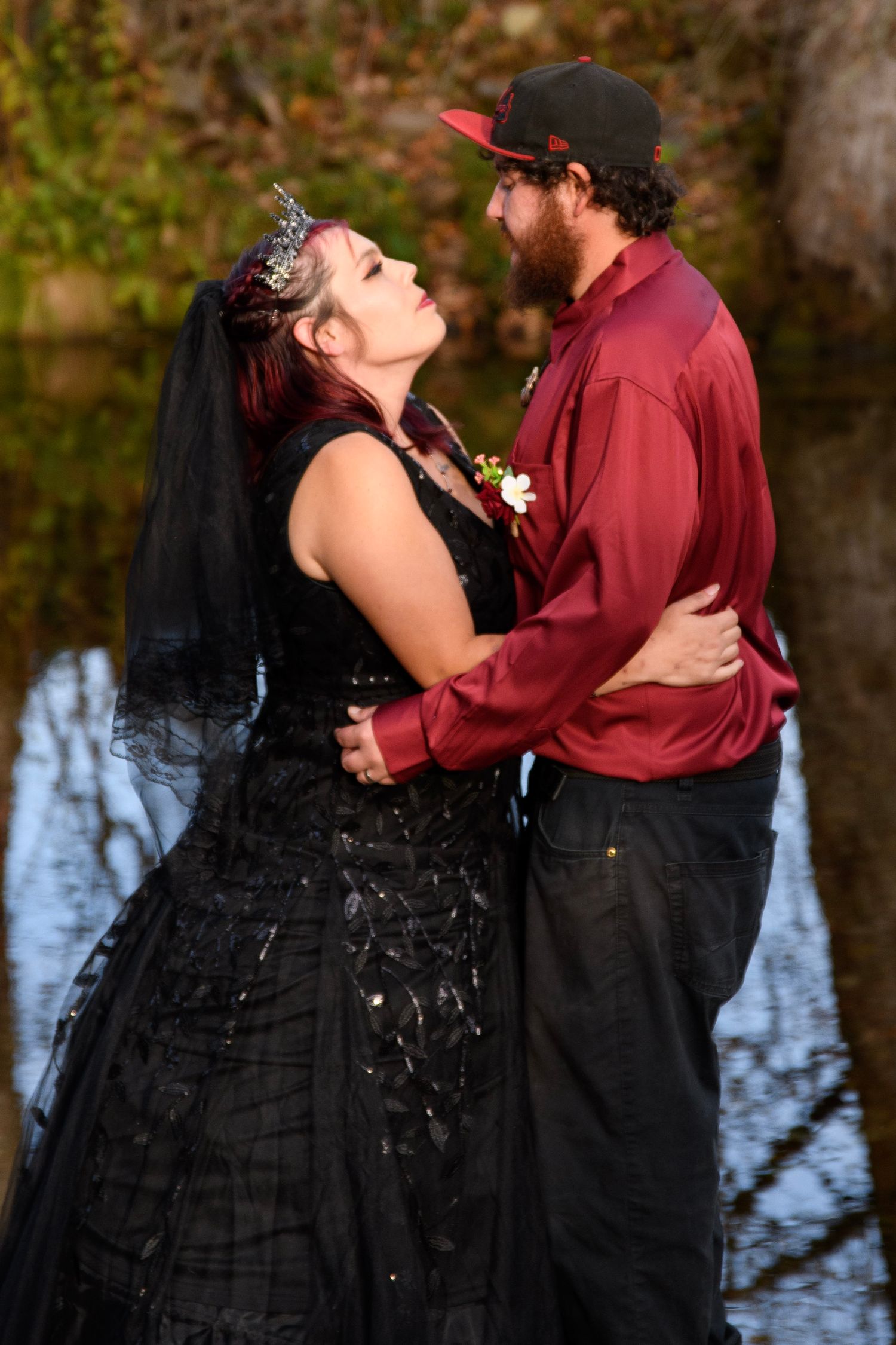 Gothic wedding couple gazing at each other, bride in black dress with veil and crown, groom in red shirt outdoors.