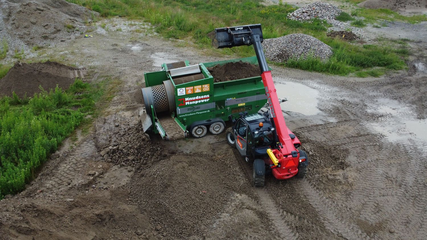 Green industrial shredder machine processing soil and debris at an outdoor construction site on a cloudy day.