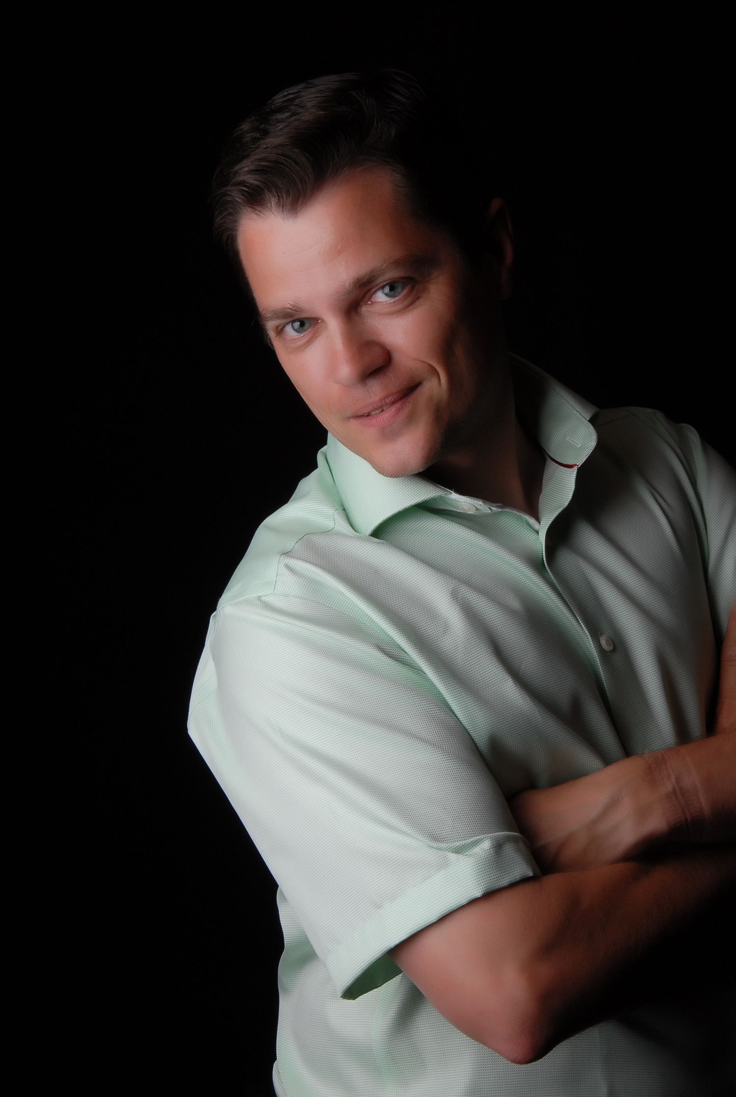 Confident man in light green shirt with arms crossed, smiling against dark background in professional portrait.