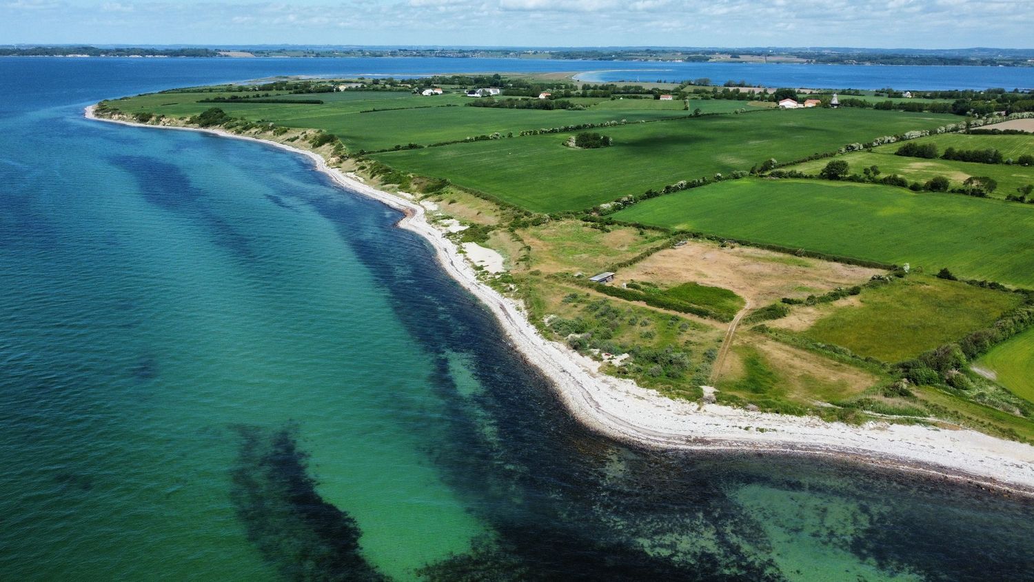 Aerial view of a scenic coastal peninsula with turquoise waters, white sandy beach, and lush green farmland fields.