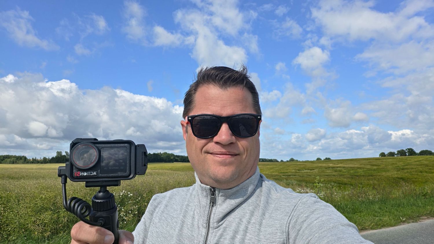 Man in sunglasses takes a selfie with an action camera on a handheld grip in a sunny green field.