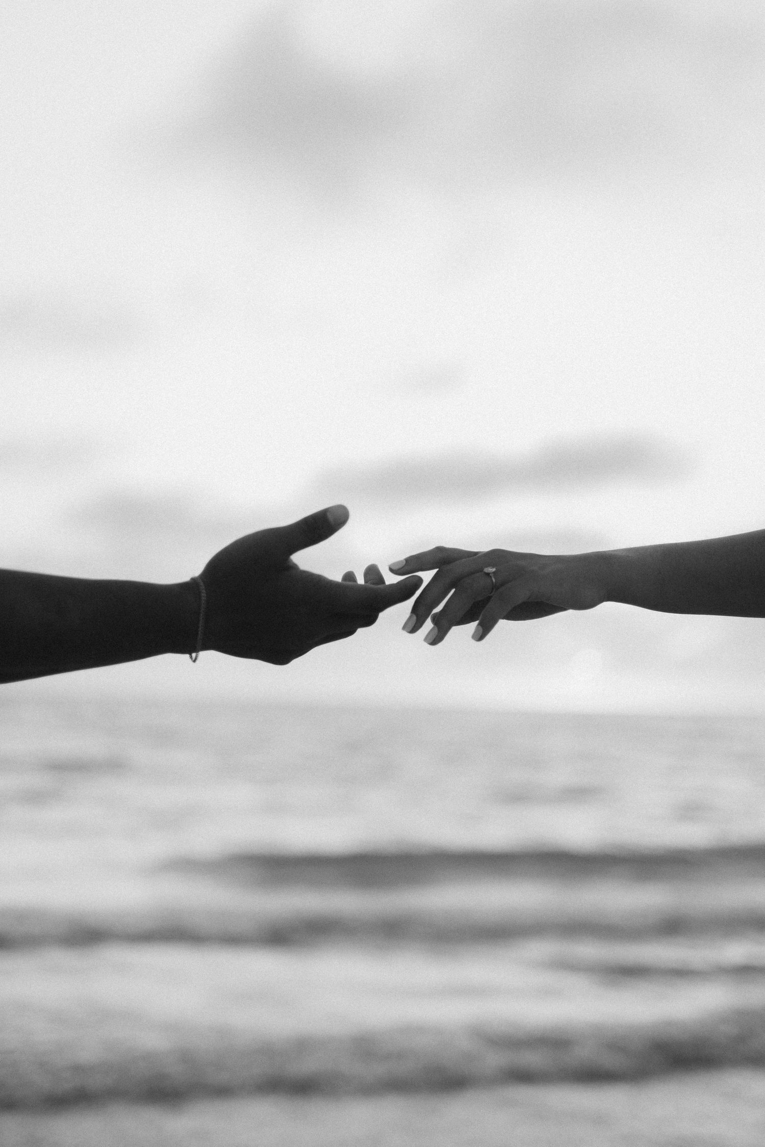 A black and white image of two hands reaching for each other in front of the ocean. One hand has a diamond ring.