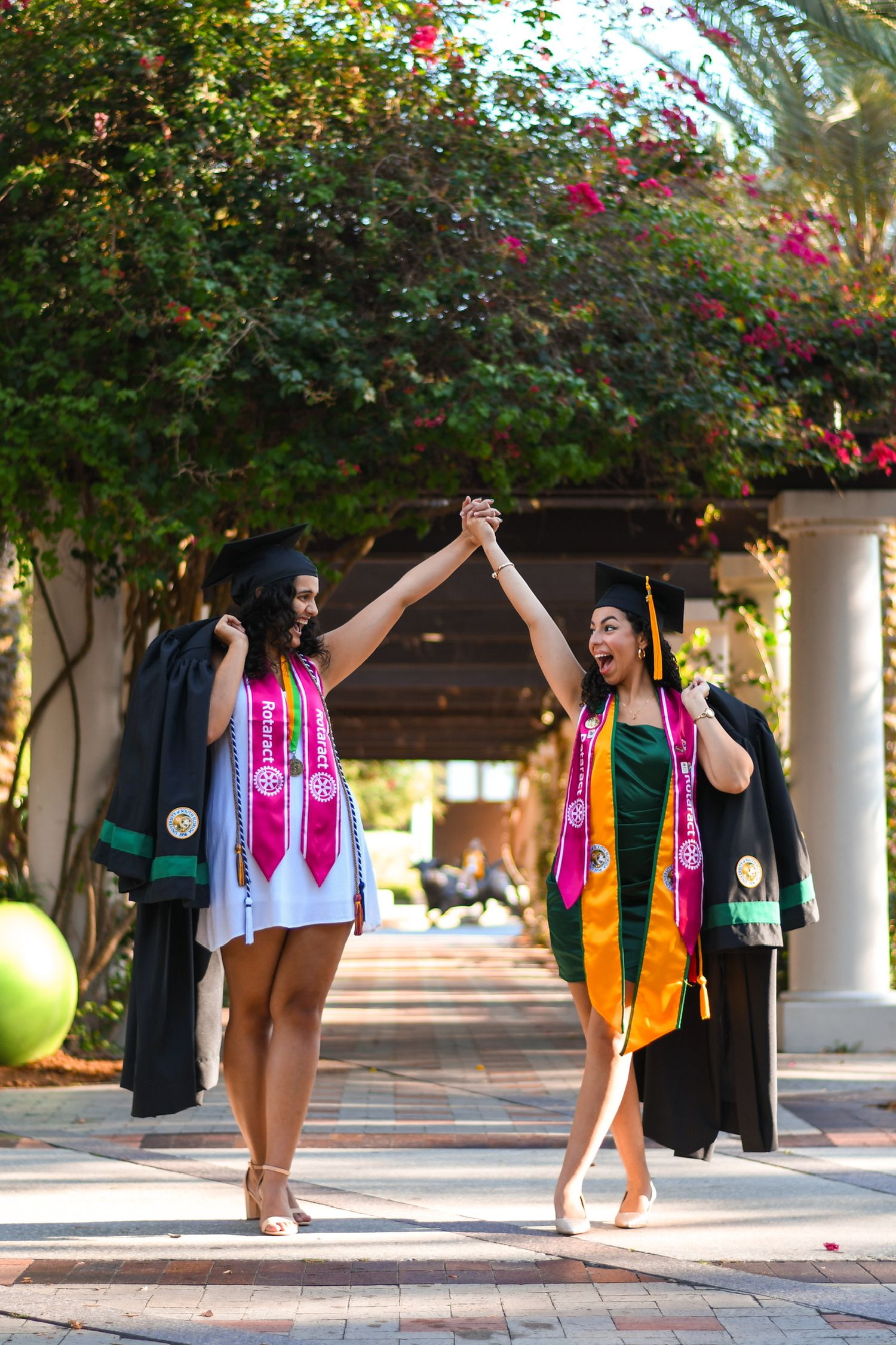 2 women in graduation regalia holding hands and looking at each other in celebration.