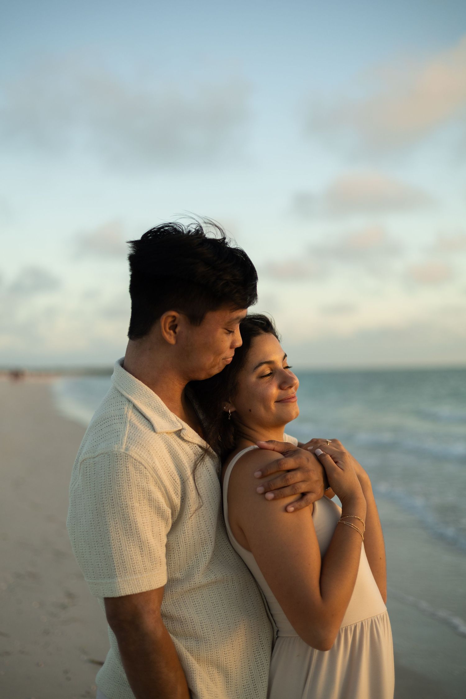 A man in a white shirt smiling down at a woman in a white dress he is embracing as they stand on the beach and look at the ocean.