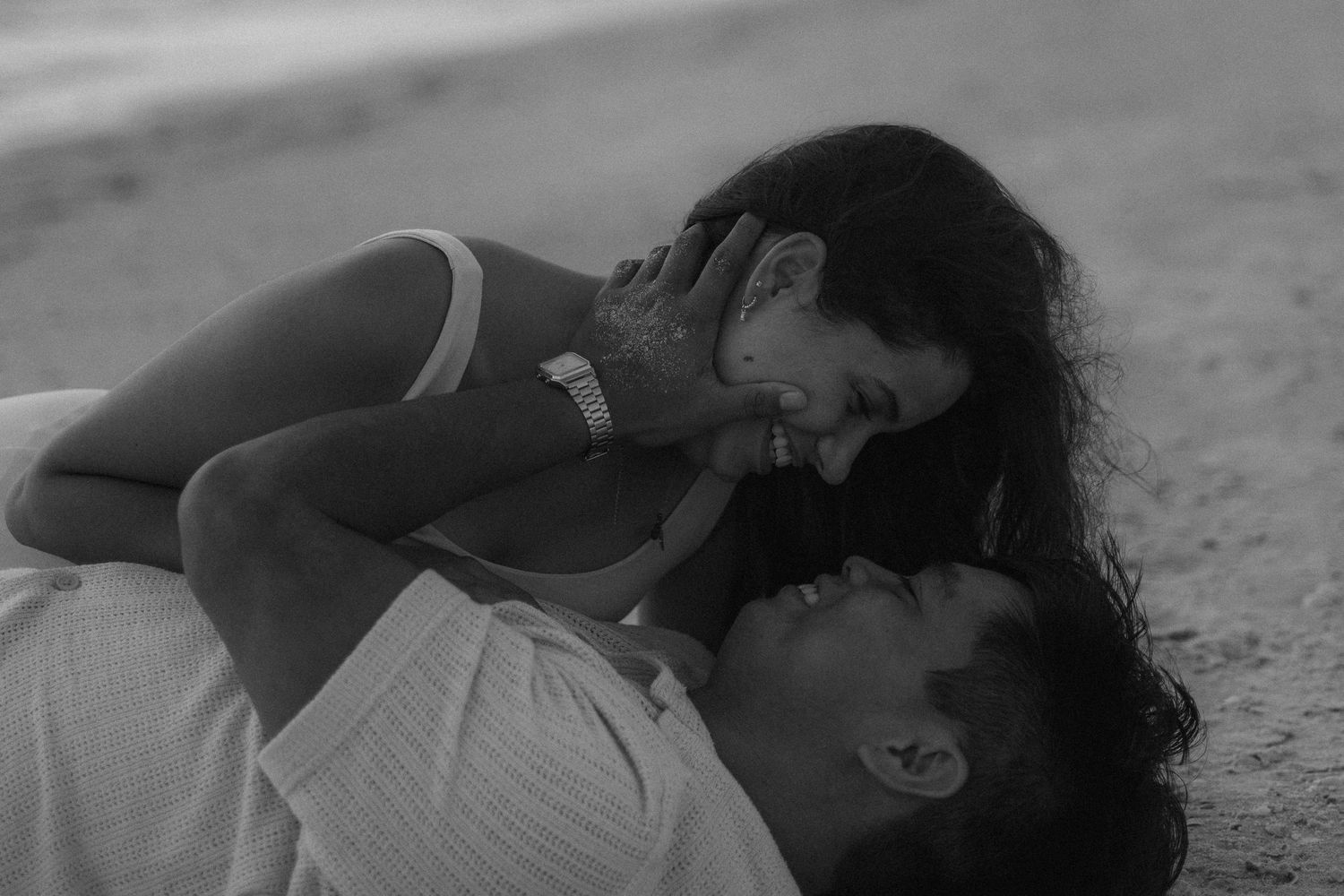 Black and white image of a man and woman laying in the sand looking at each other, both wearing white.