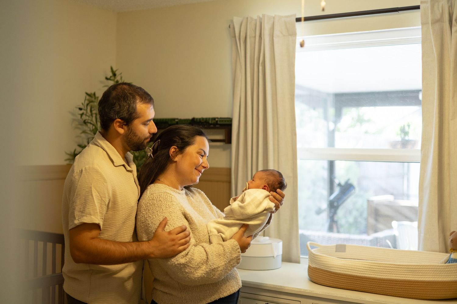 A mom and dad staring down and smiling at their newborn baby. They are all wearing white standing in the baby's nursery.