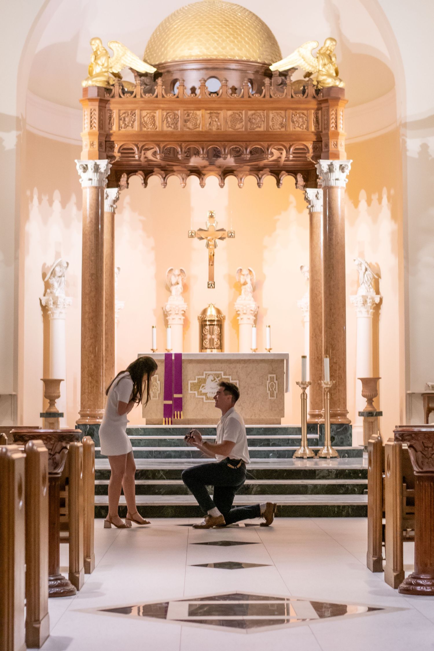 A man in a white shirt on one knee proposing to a woman in a white dress in a church.