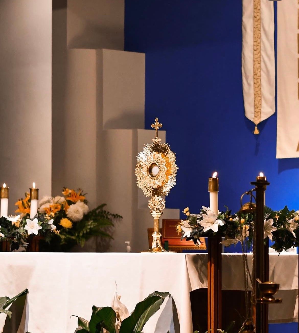 Light shining on a gold monstrance with the Eucharist inside atop an alter with candles and flowers around the alter.