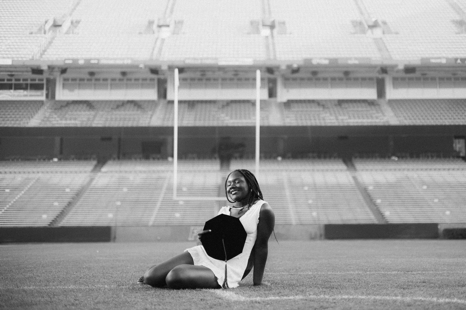 Black and white image of a woman in a white dress sitting on a football field, closing her eyes and smiling, holding a black graduation cap.