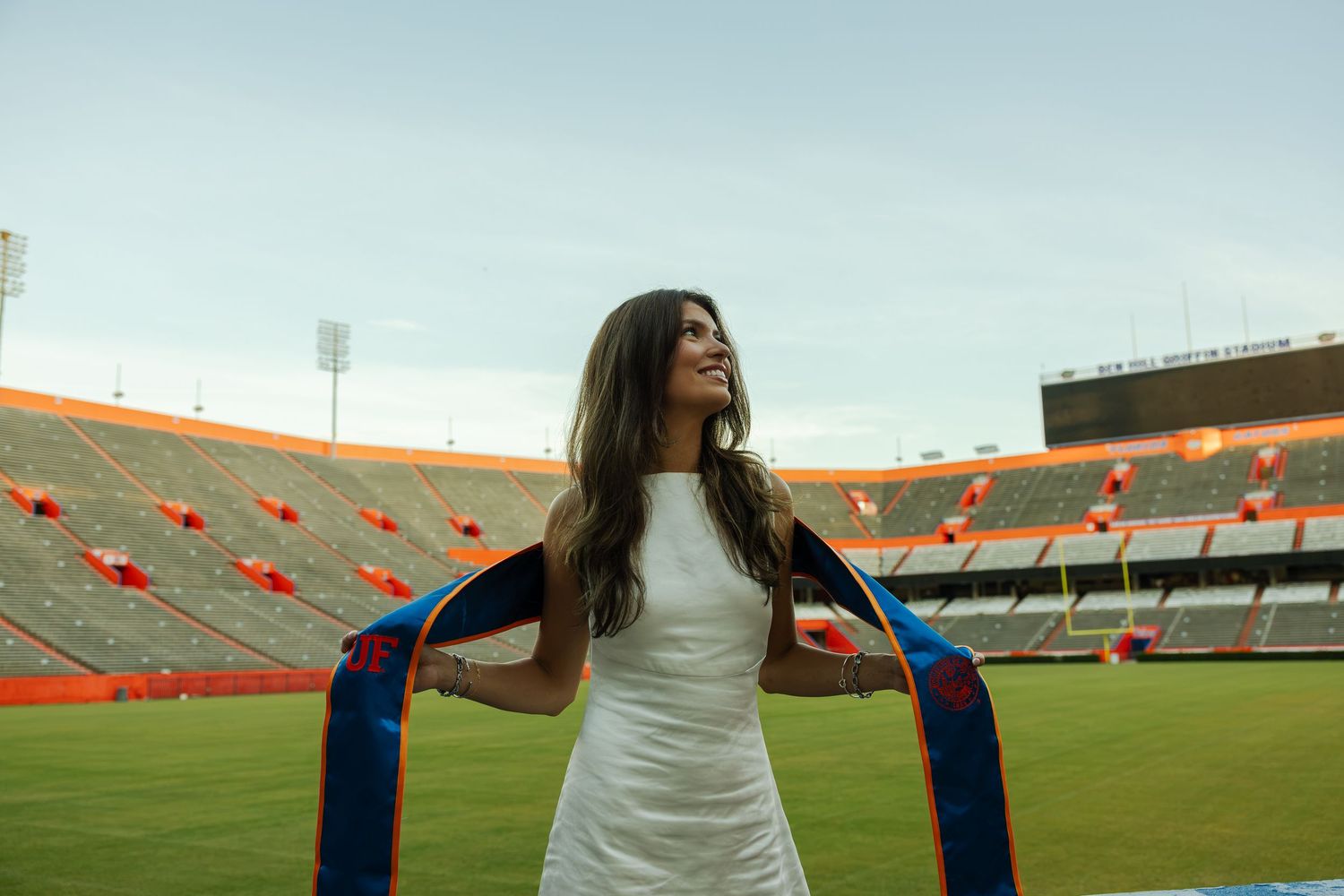 A girl in a white dress standing in front of the grass in a stadium looking up at the blue sky with a blue and orange stole around her.