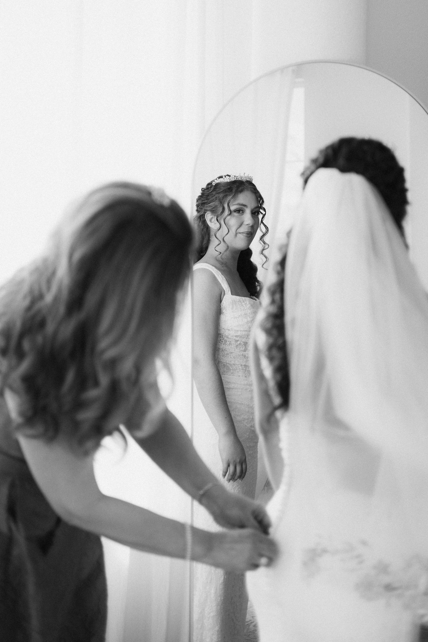 A black and white image of a woman wearing a wedding dress looking at herself in a mirror while another woman buttons up her dress.