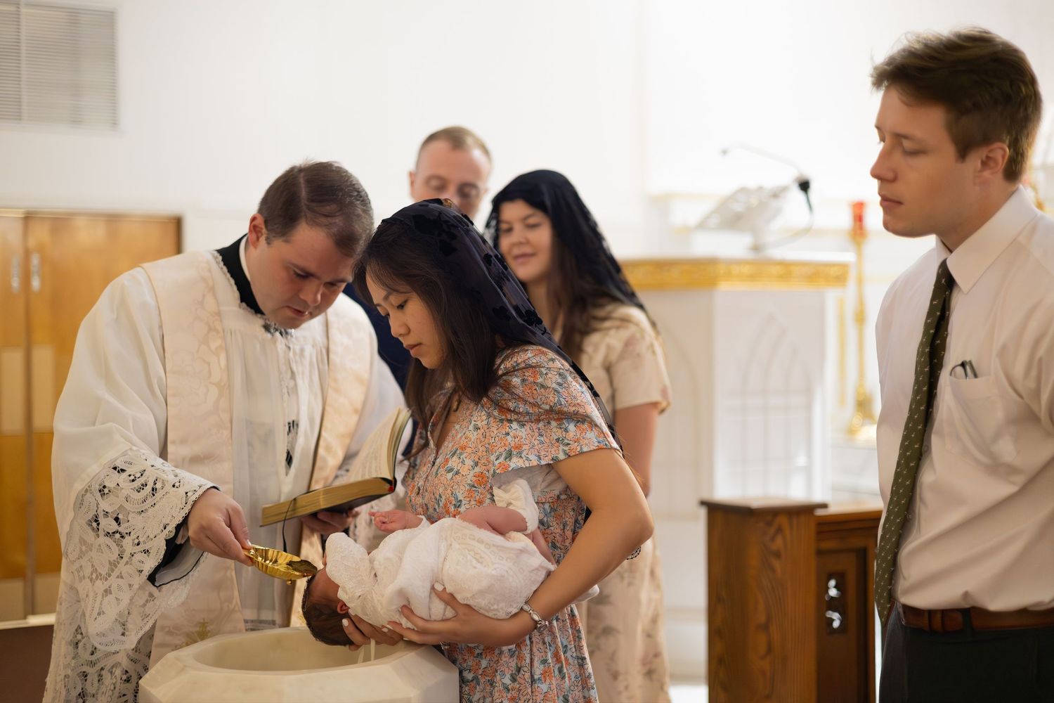 A woman holding a baby as a priest pours water over the babies head. A woman and two men watch smiling.