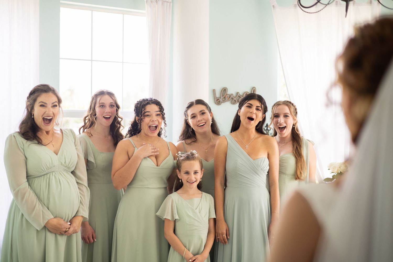 Six women and one girl in light green dresses gasp and smile at a woman in a wedding dress.