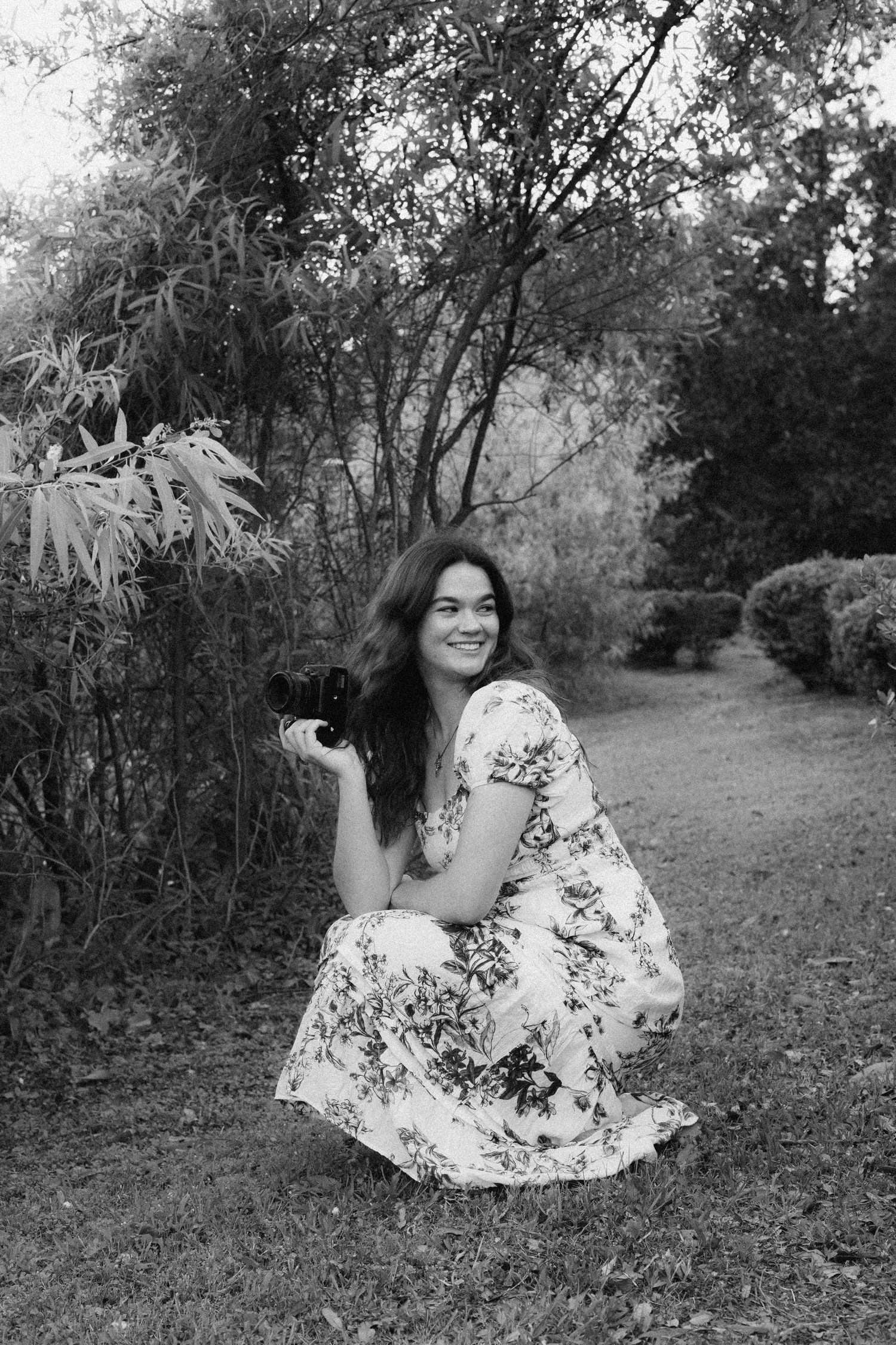 A black and white image of a woman with long dark hair wearing a long dress squatting while holding a camera.