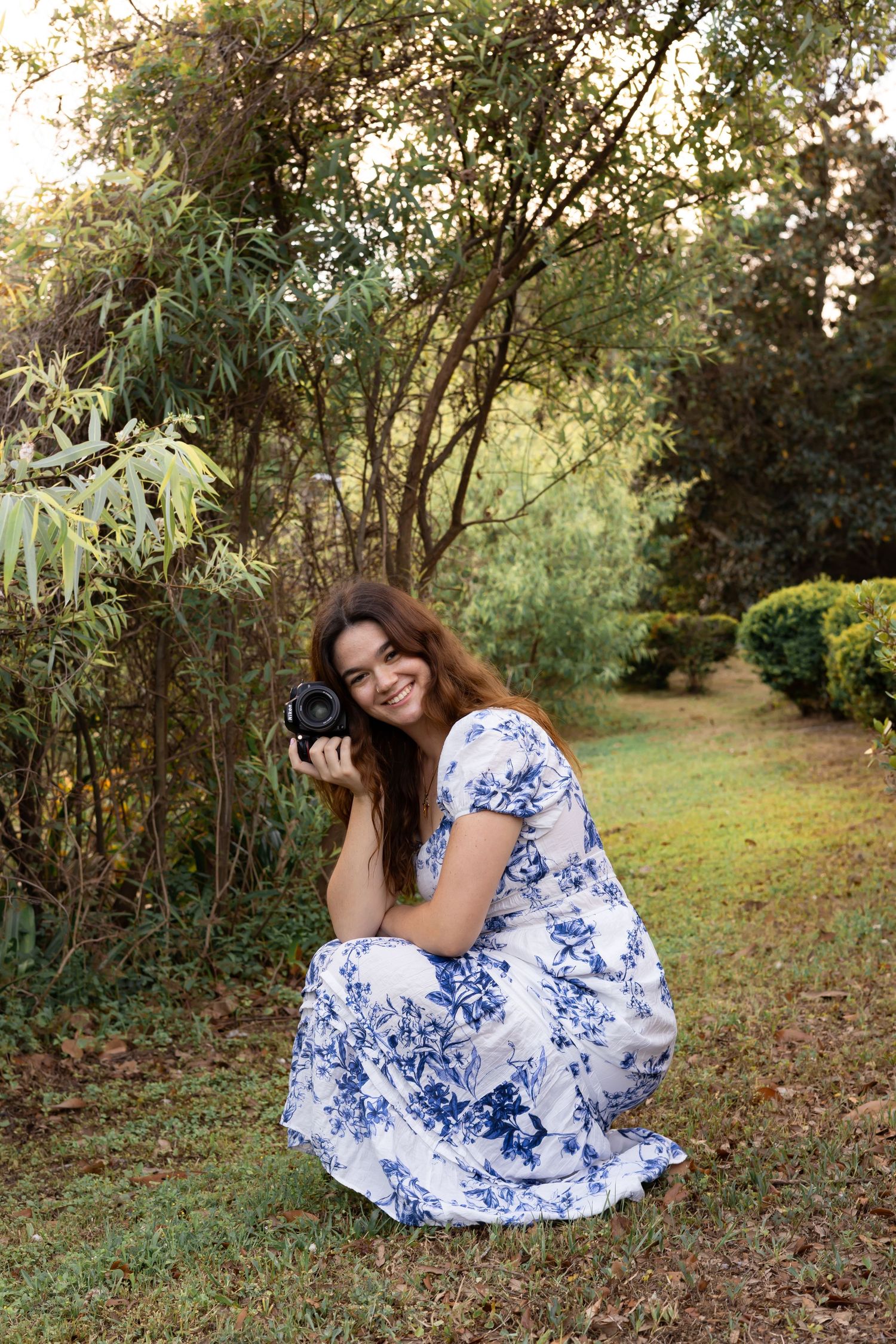 A woman squatting in a long blue and and white dress with greenery behind her holding a camera and smiling.