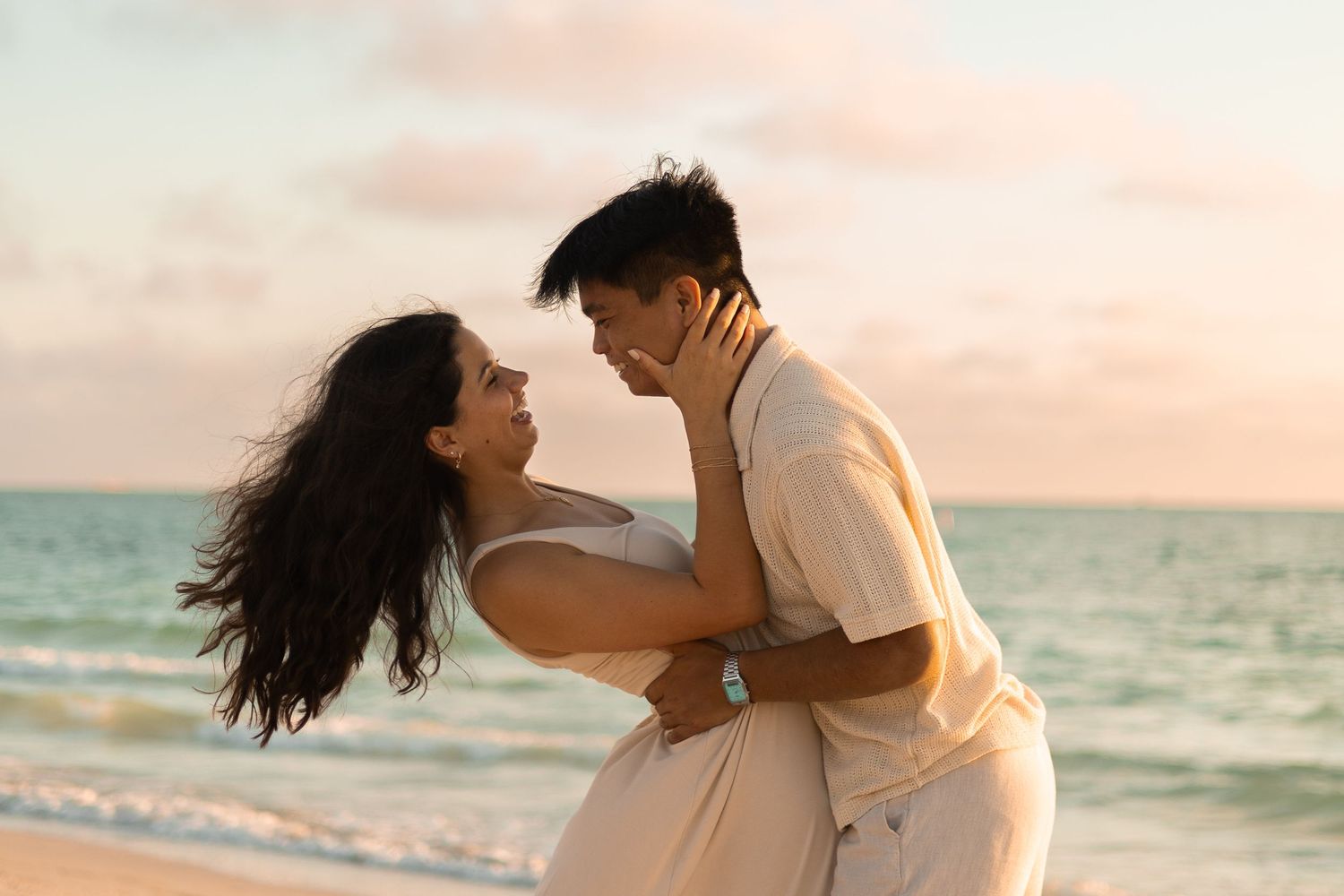 A man and woman both wearing white smiling at each other on the beach. Her hands are on his face and his hands are on her waist.
