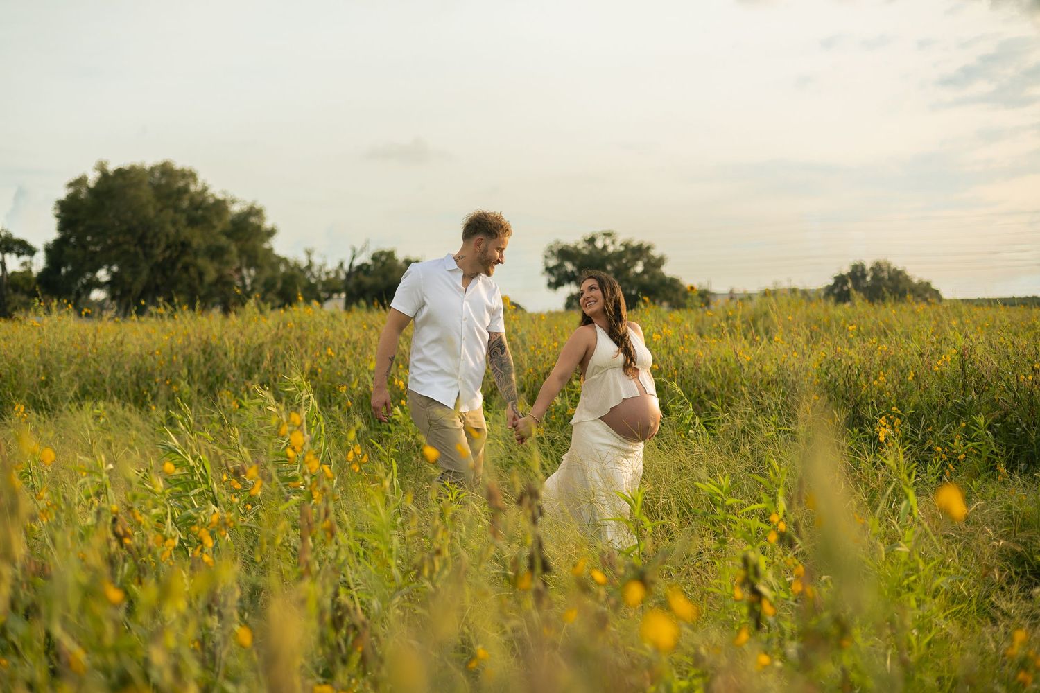 A man and a pregnant woman wearing white walking through and field and smiling.