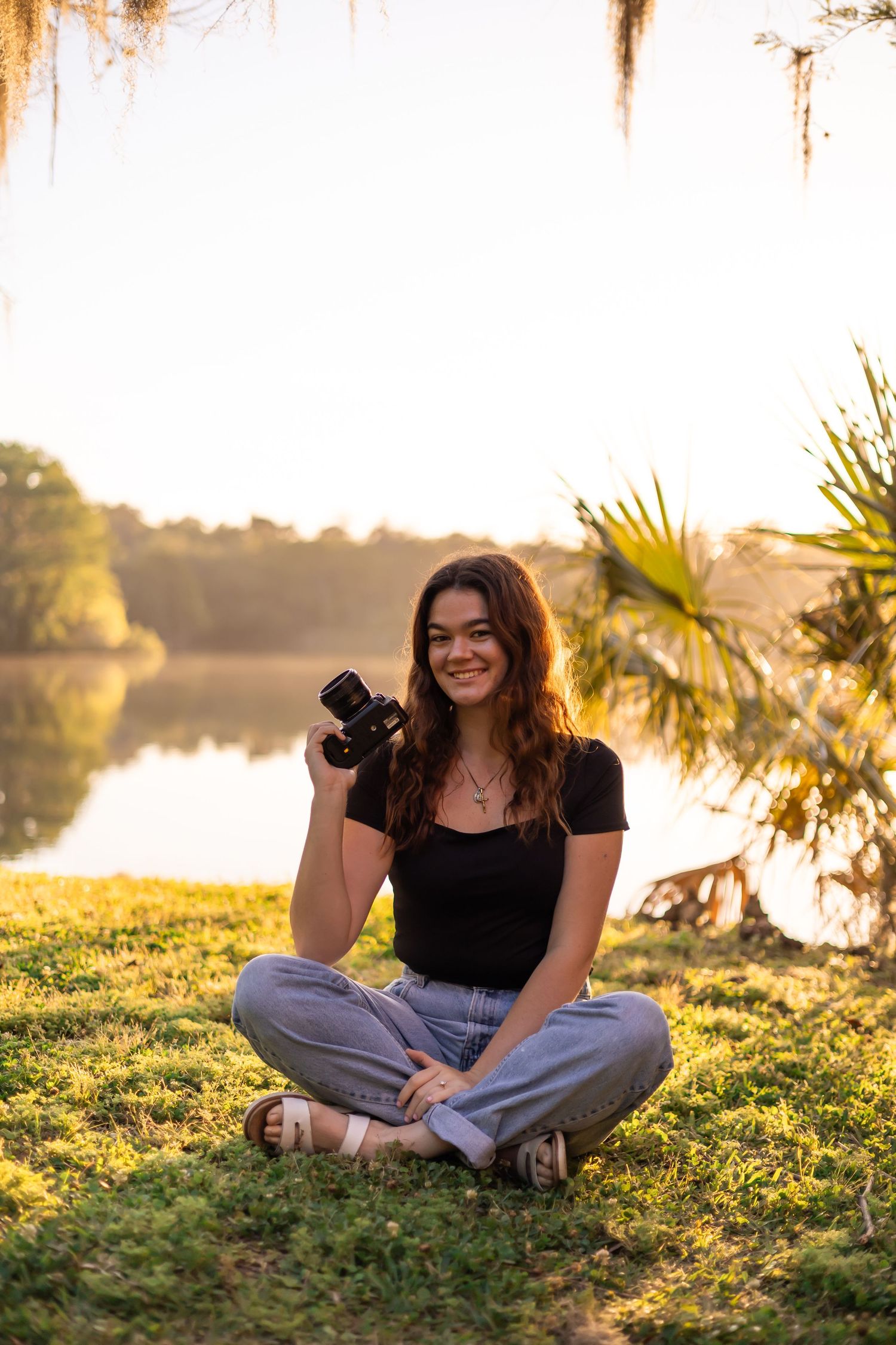 A woman in a black shirt and jeans sitting in grass holding a camera and smiling.