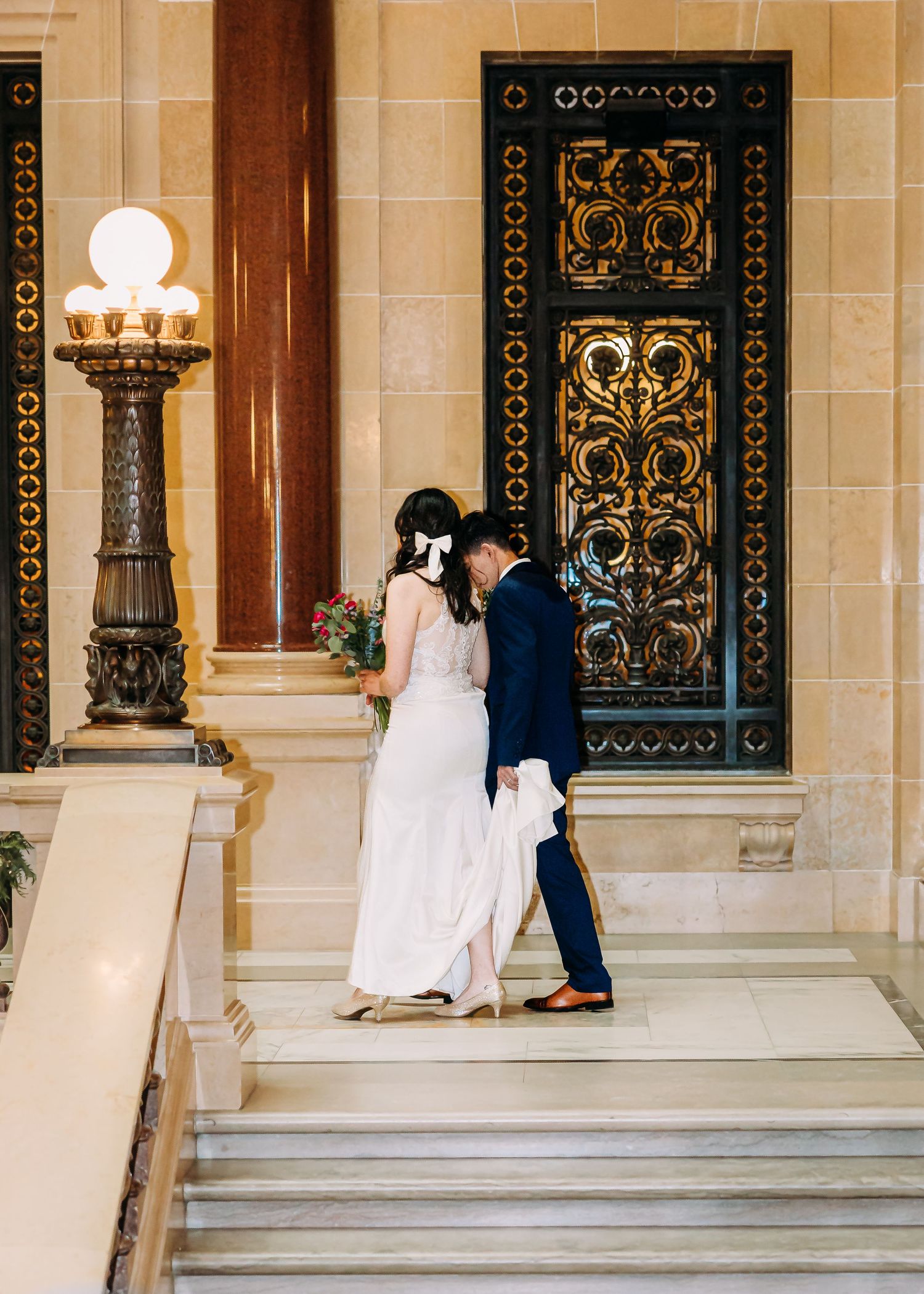 Bride and groom share a tender moment on marble staircase beside ornate gilded door.