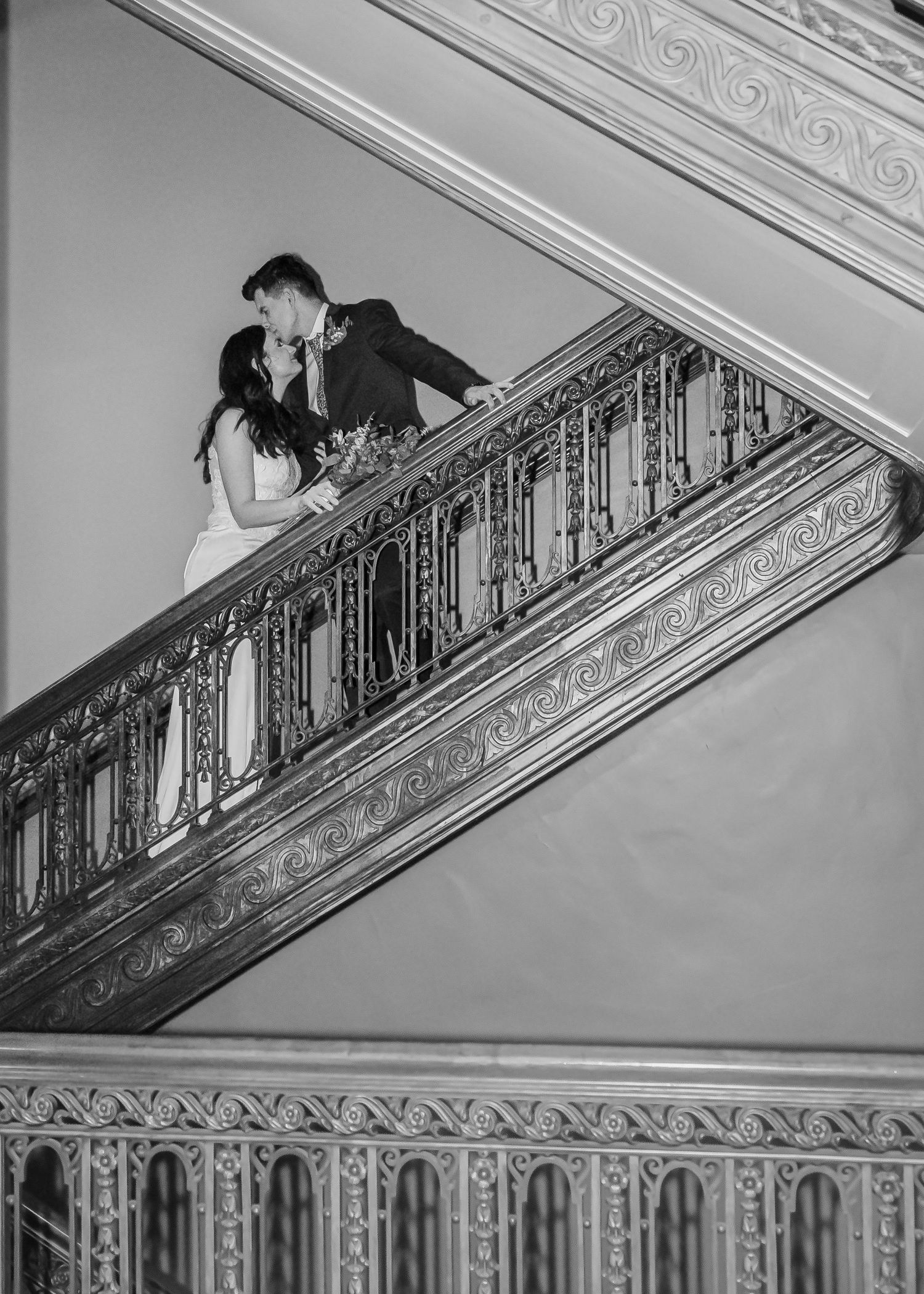 Black and white photo of bride and groom kissing on an ornate wrought iron staircase railing.