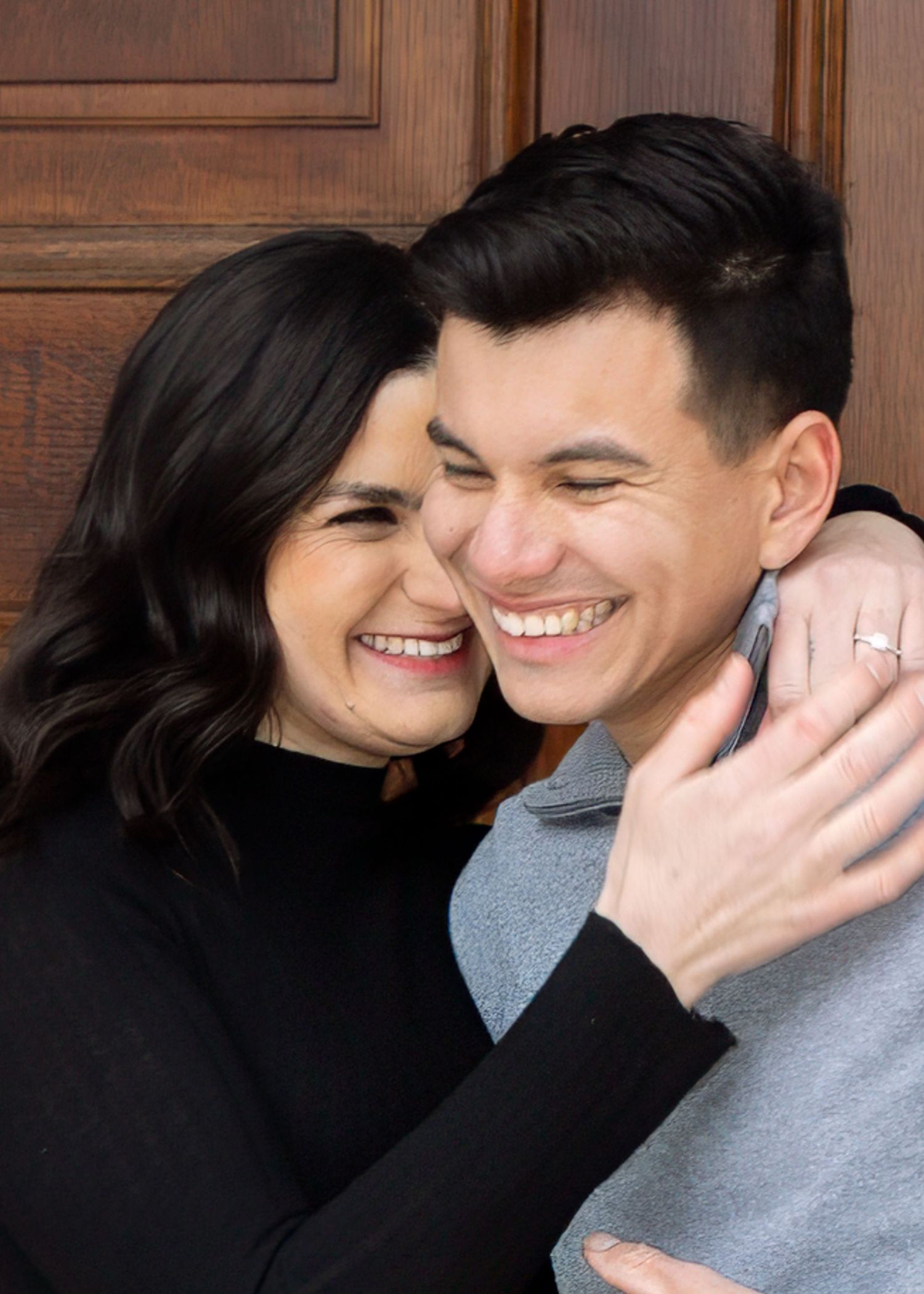 Happy couple laughing and hugging closely in front of a wooden door, woman wearing engagement ring.