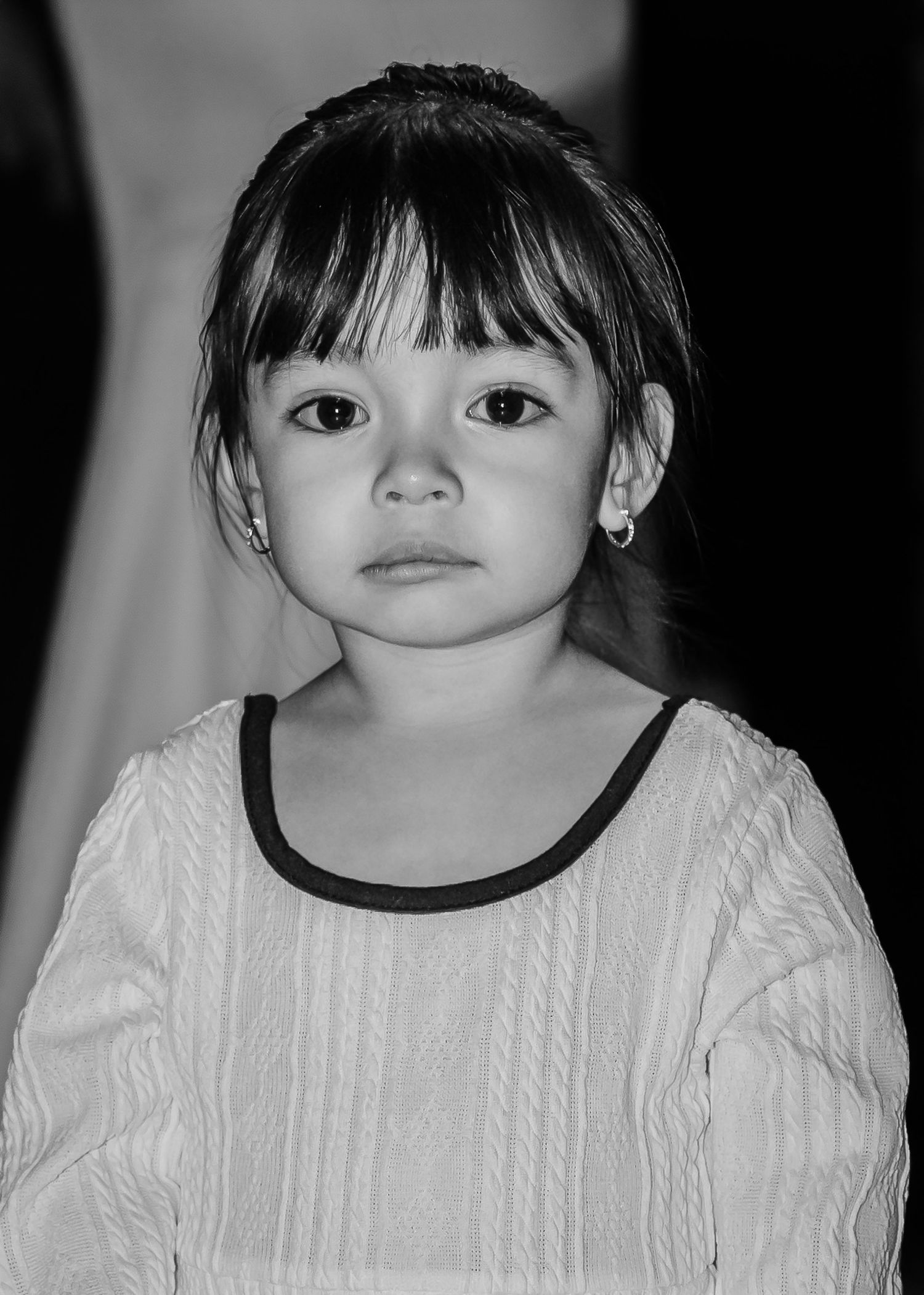 Black and white portrait of a young girl with bangs wearing a white sweater and small earrings.