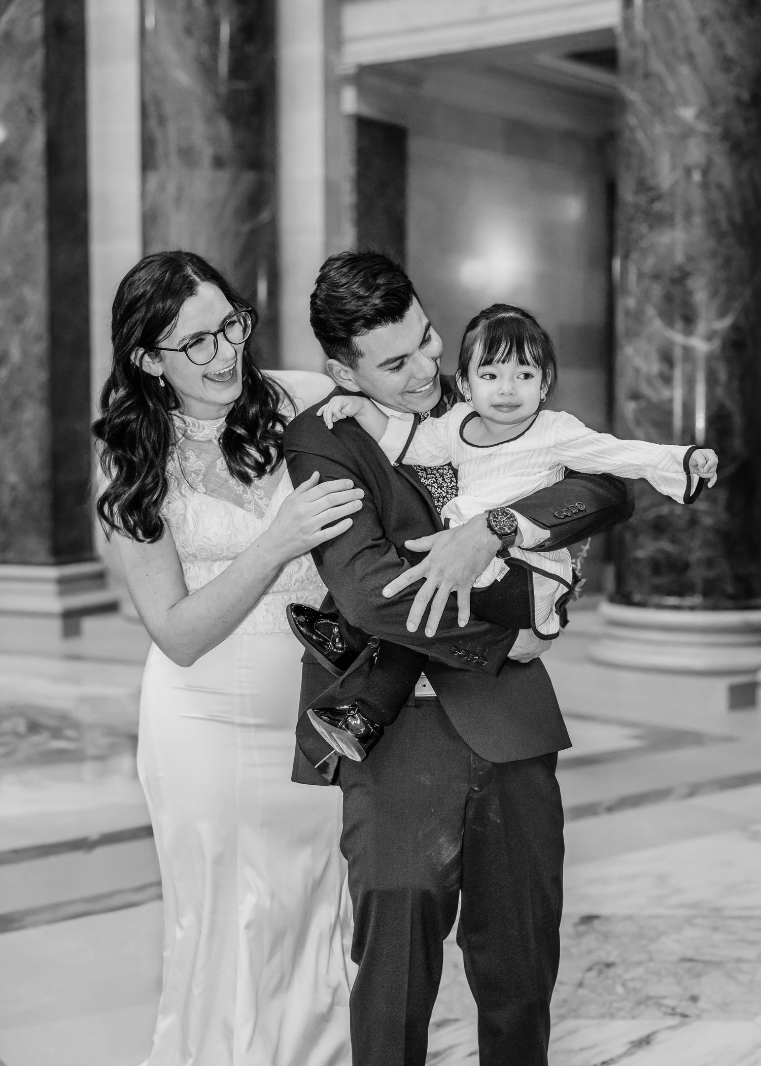 Black and white photo of newlyweds in elegant venue holding a happy toddler between them.