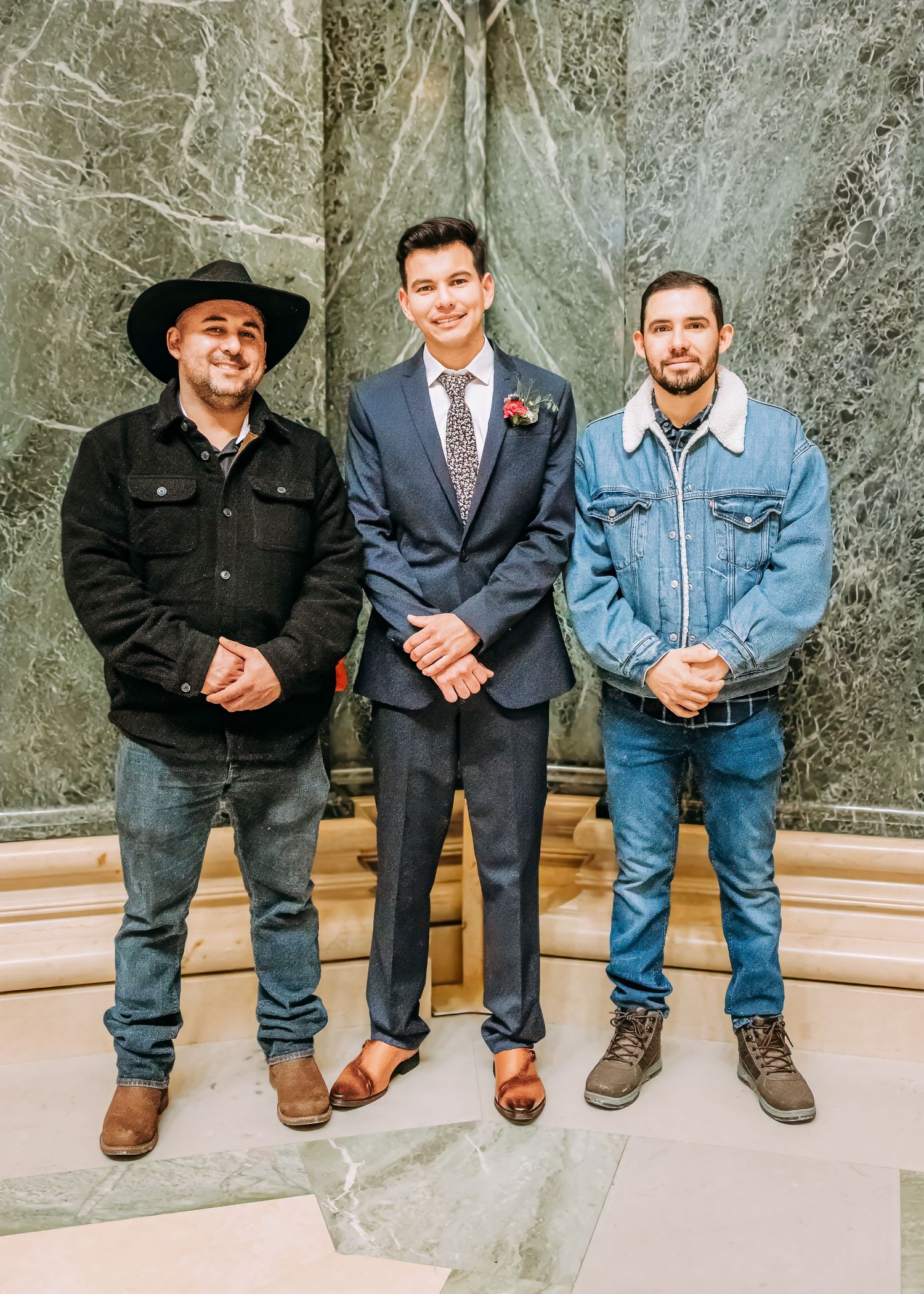 Groom in navy suit smiling with two casually dressed guests in a marble-columned wedding venue.