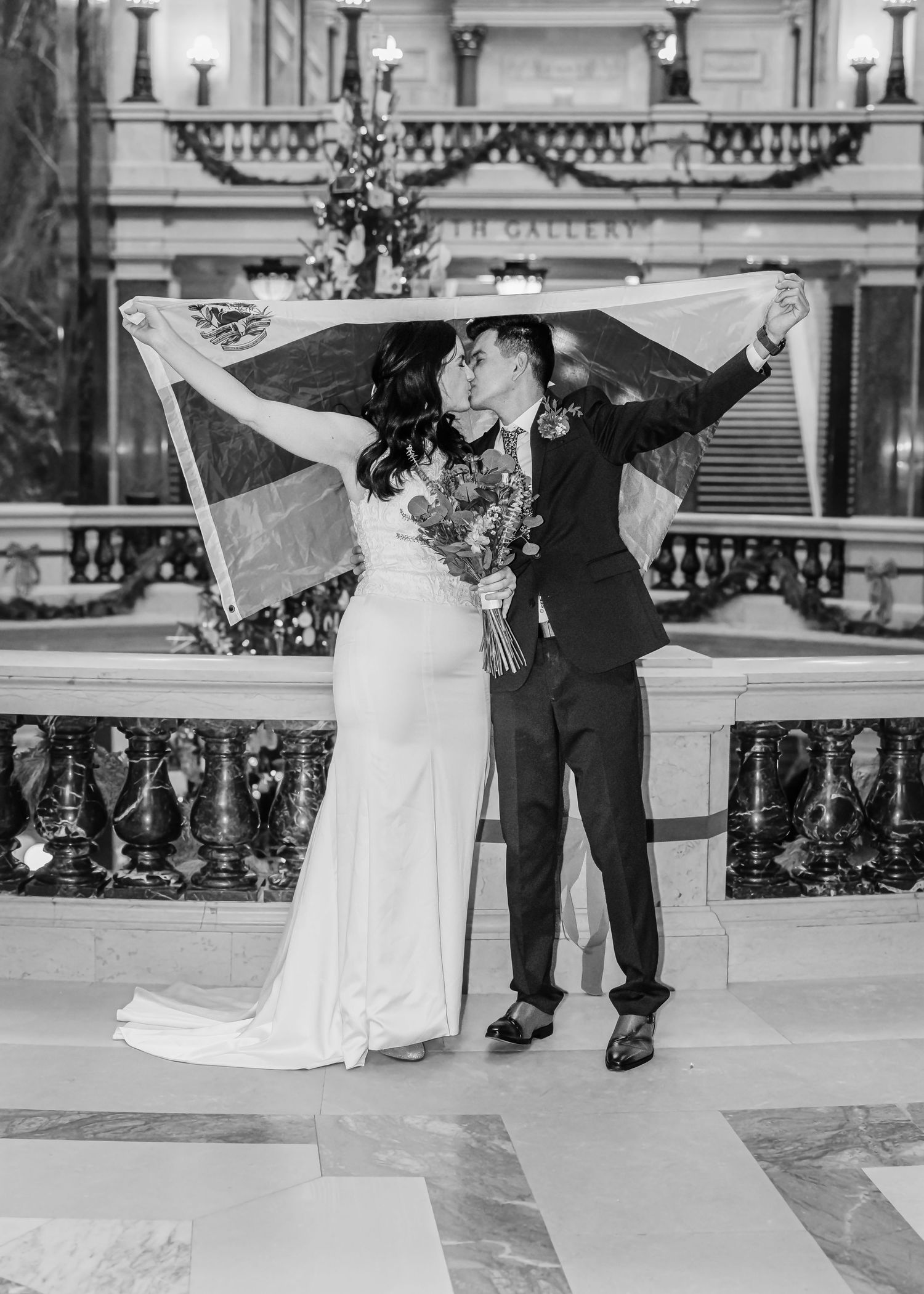 Bride and groom share a kiss while raising a flag in a grand illuminated hall.