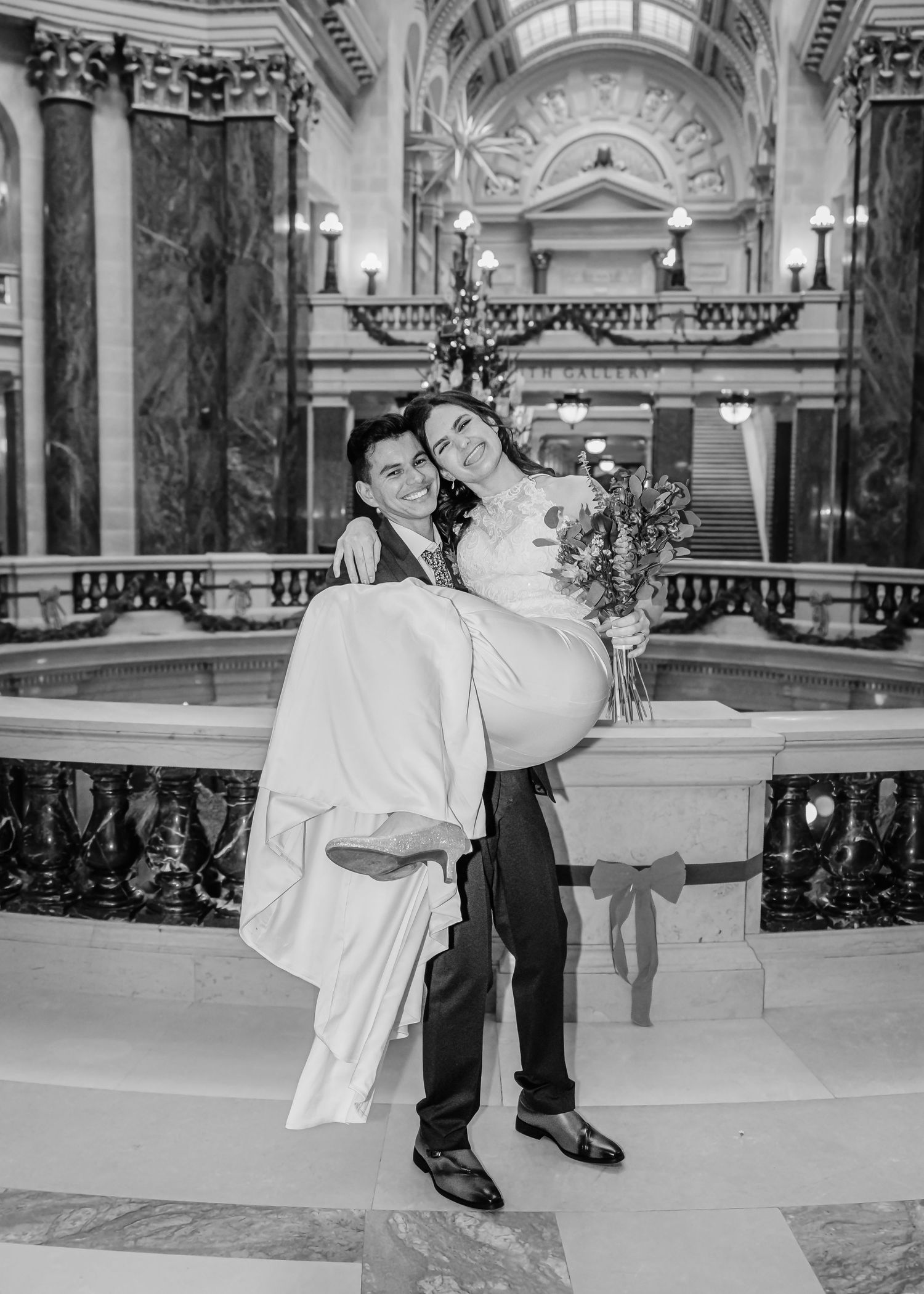 Groom carries smiling bride in elegant black and white wedding photo inside grand marble rotunda.