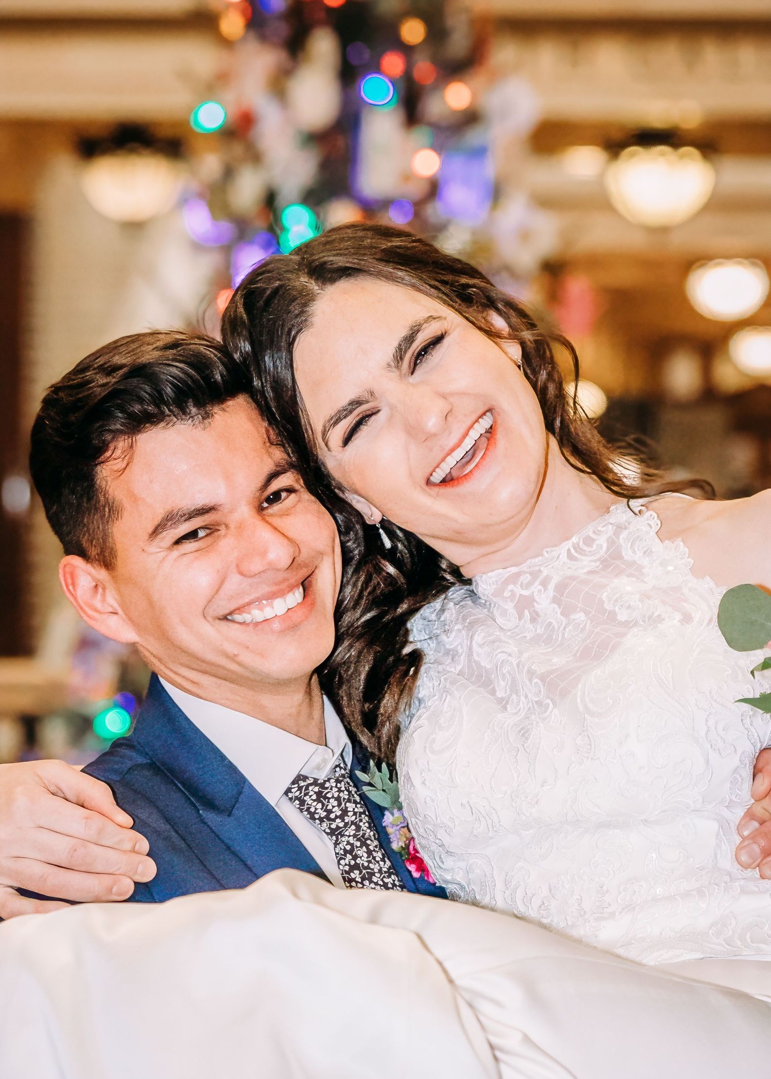 Joyful bride and groom embracing and laughing together in front of a decorated Christmas tree at their wedding.