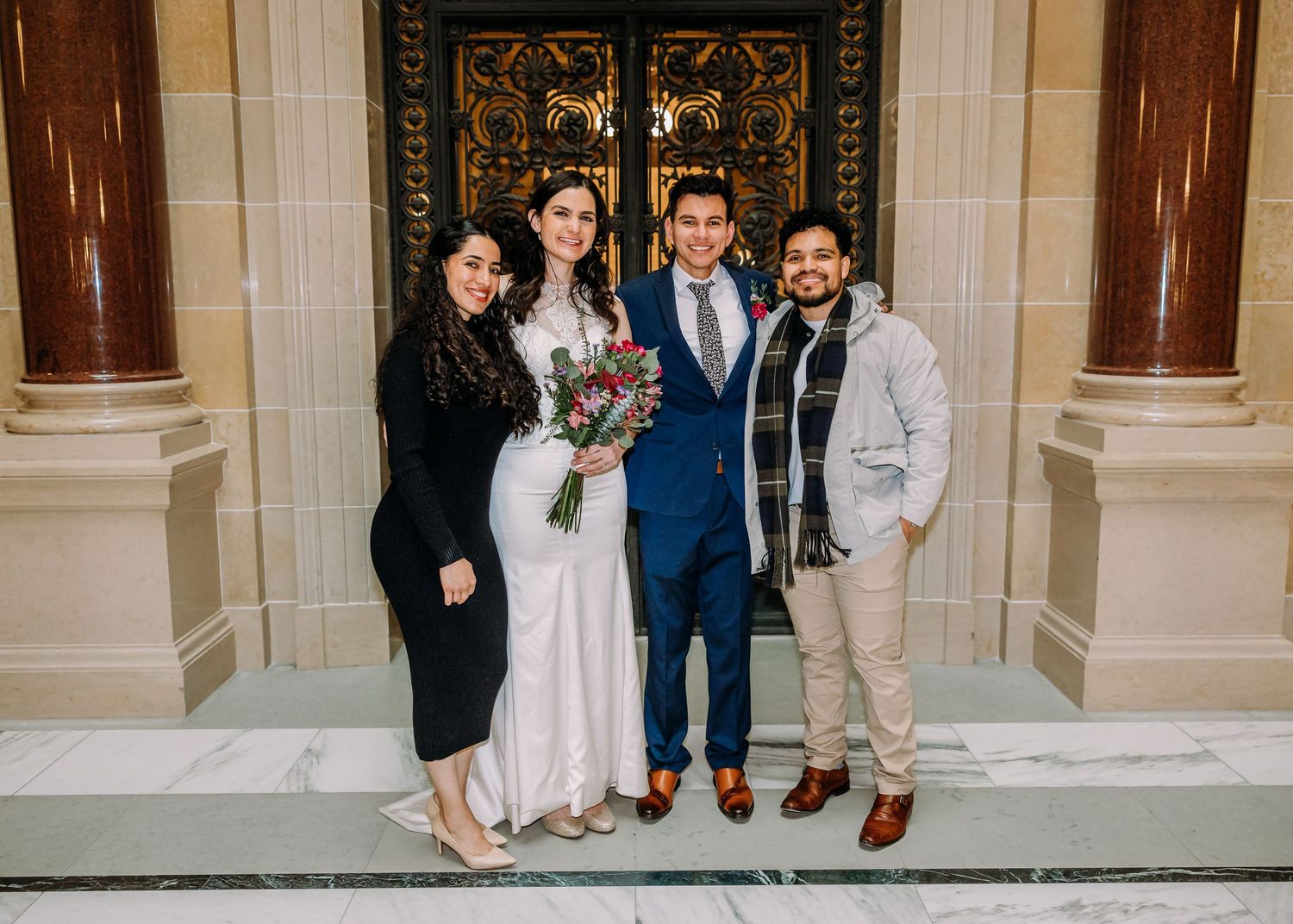 Wedding couple and two guests smile together before decorative golden doors at a formal city hall ceremony.
