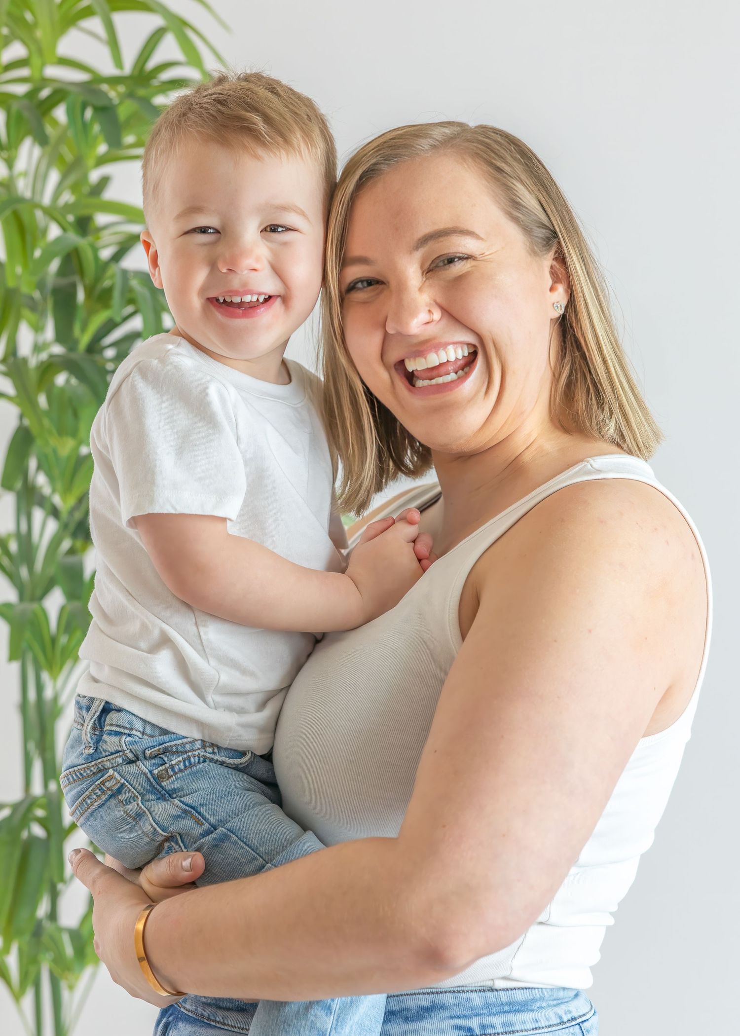 Laughing blonde mother and smiling toddler son sharing joyful moment in bright indoor portrait.