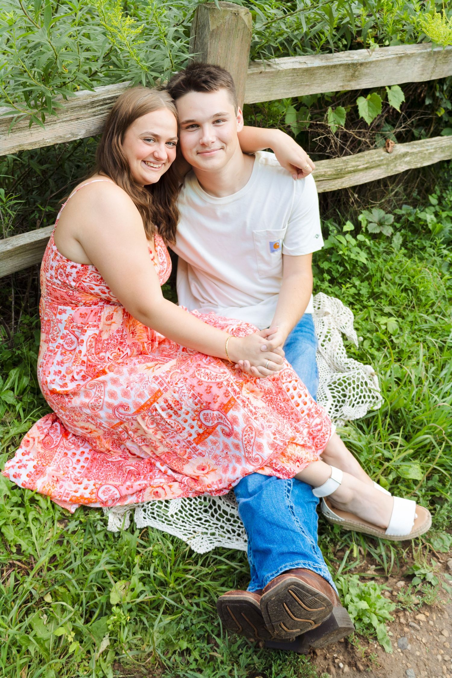 Smiling couple sitting together against a wooden fence surrounded by lush green foliage.