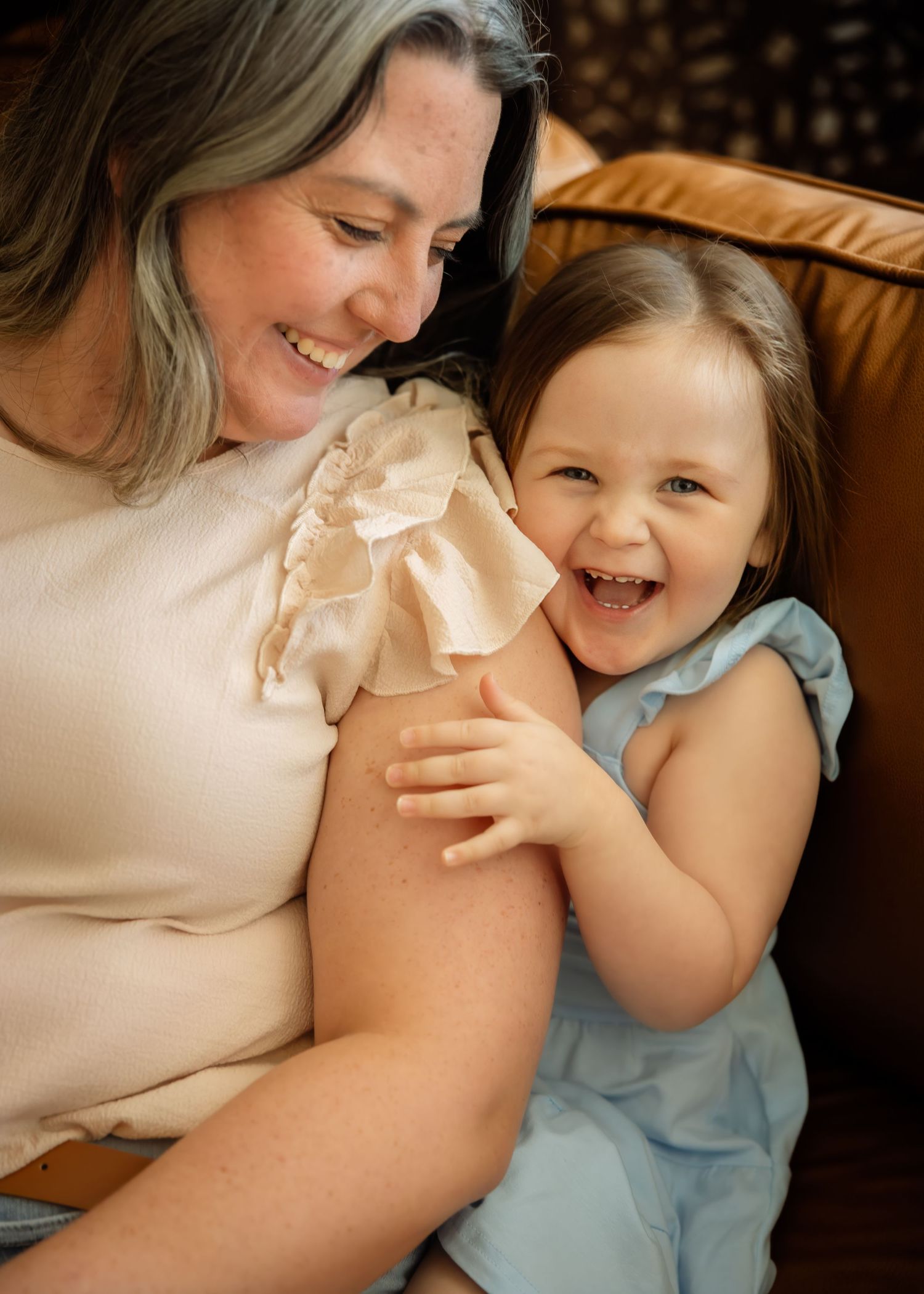 A smiling grandmother holds a laughing toddler girl in a blue dress on a leather sofa.