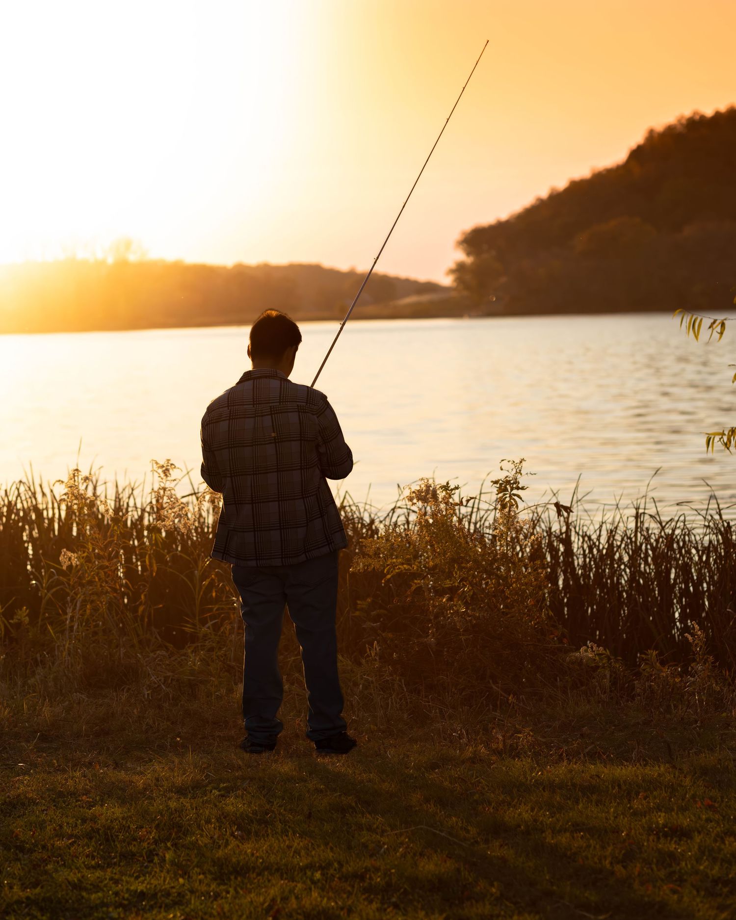 Silhouette of teen boy fishing at sunset by a peaceful lake.