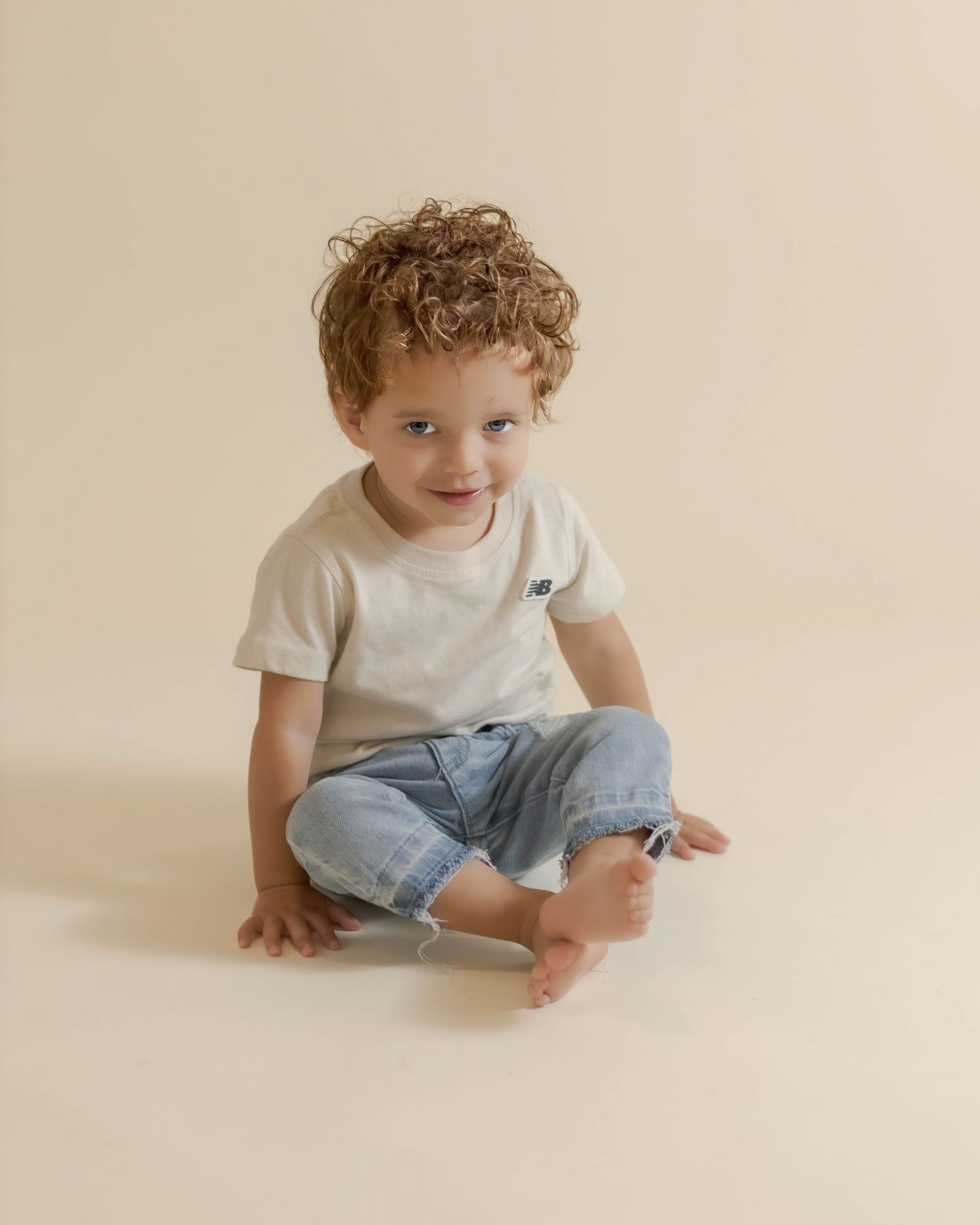 Curly-haired toddler boy sitting cross-legged in light jeans and white New Balance tee on beige background.