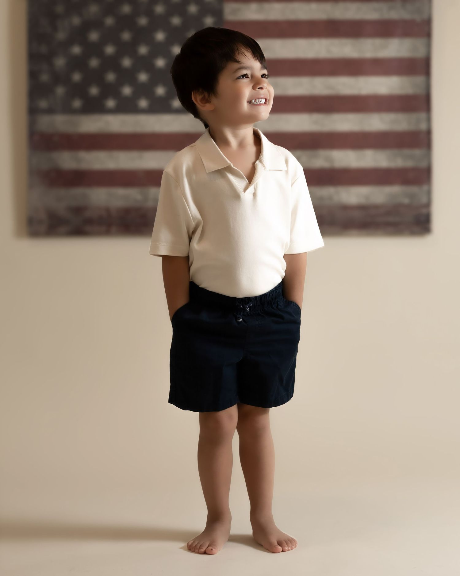 Young boy in cream polo and navy shorts smiles while standing before an American flag tapestry.