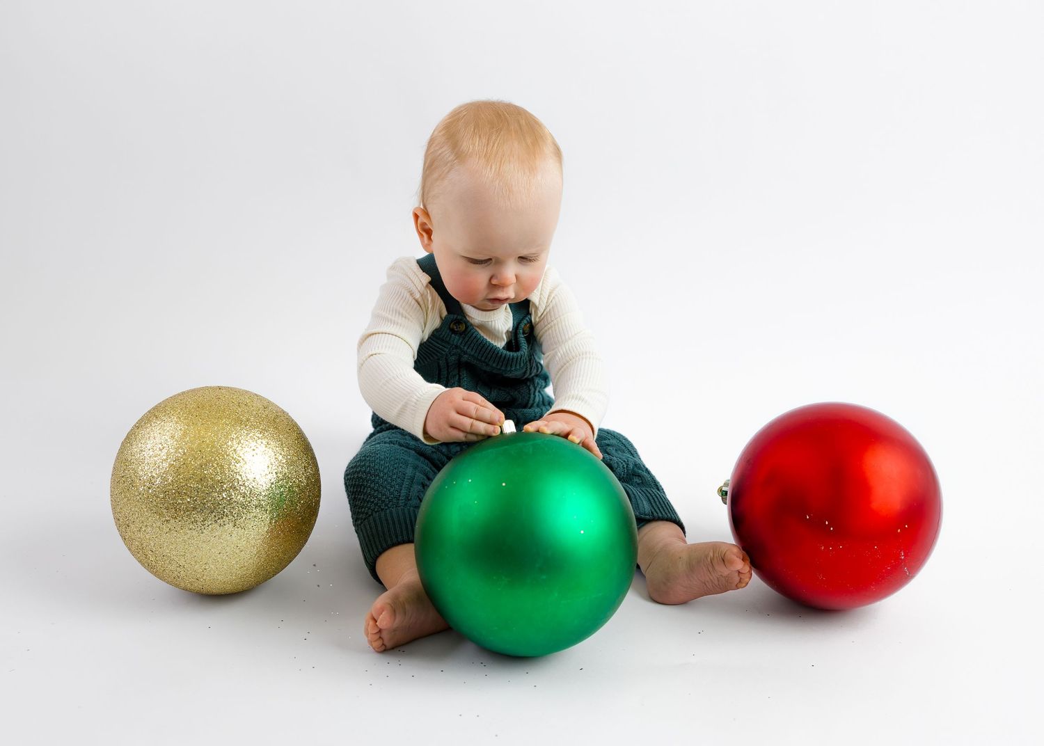 Baby in green overalls sits with large Christmas ornaments in gold, green, and red.