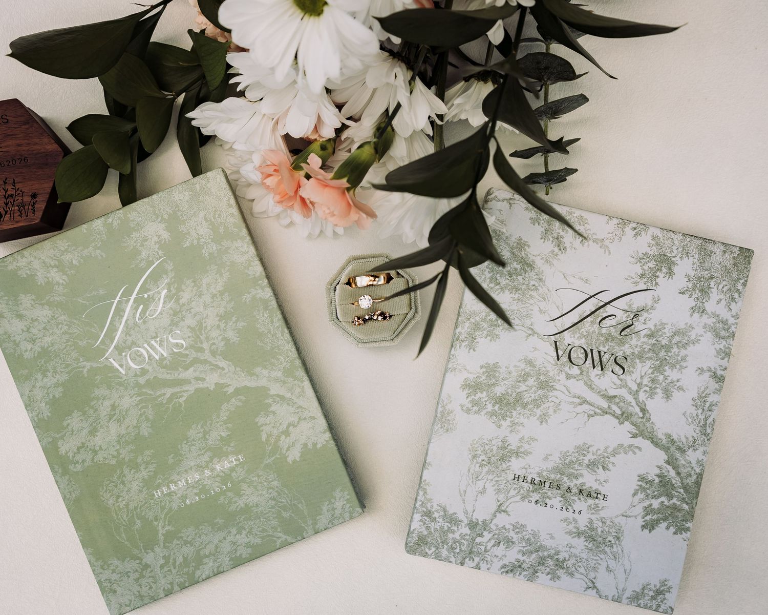 Two elegant wedding vow books in sage green and white toile designs, displayed with rings and fresh flowers.