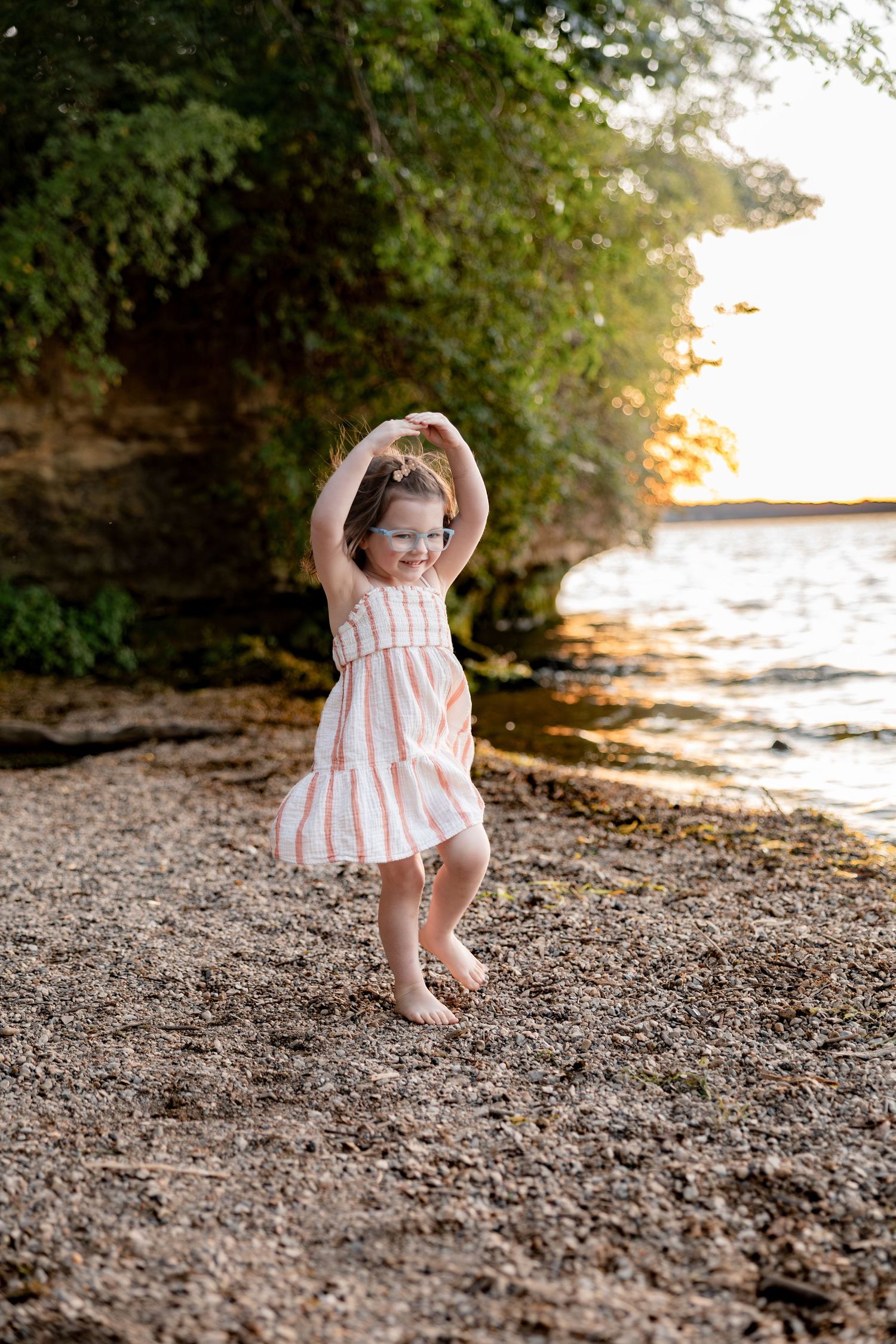 A joyful toddler girl in a striped dress dances barefoot on a rocky beach shore at golden sunset.