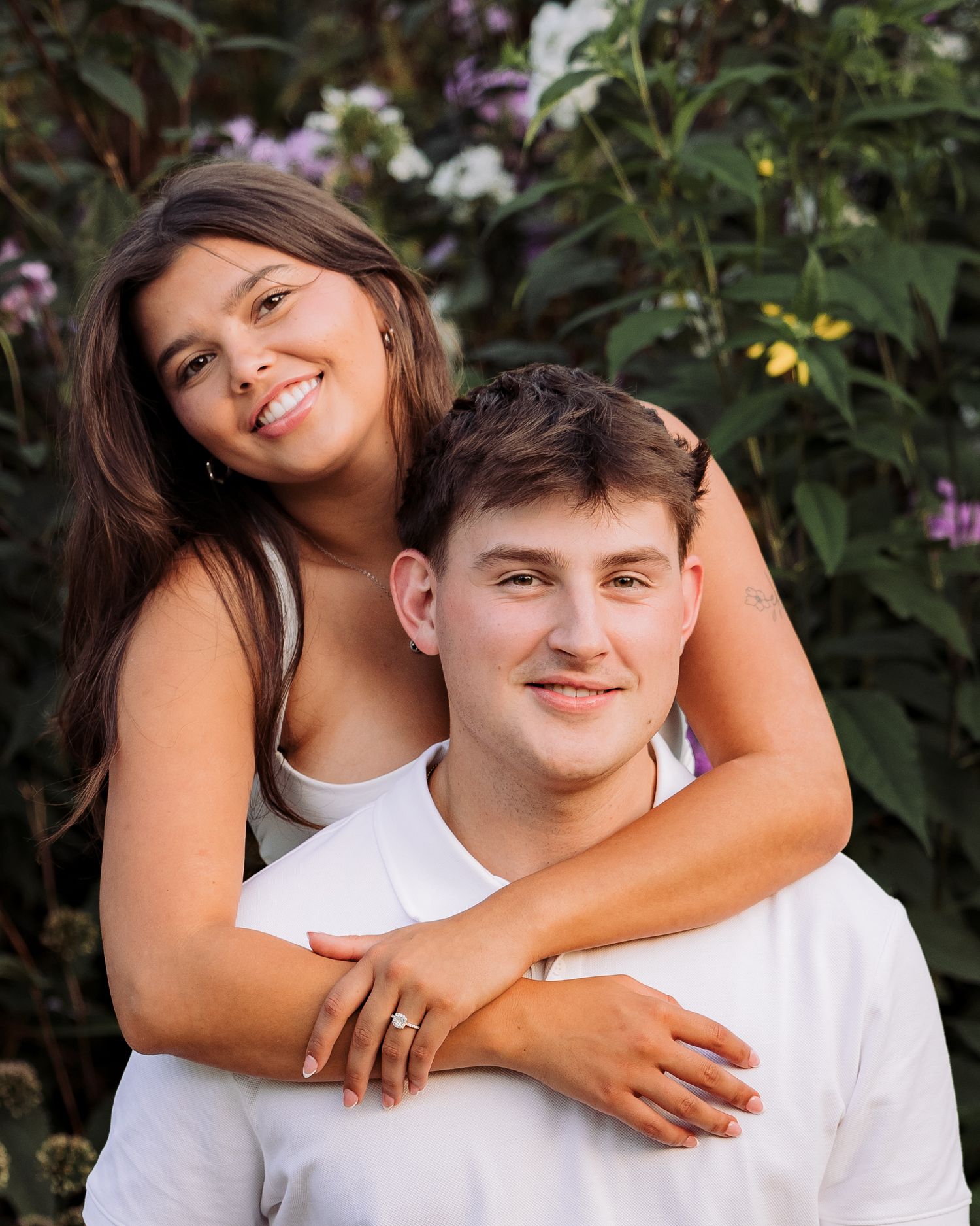 A young couple poses together outdoors, smiling in front of lush green foliage and purple flowers.