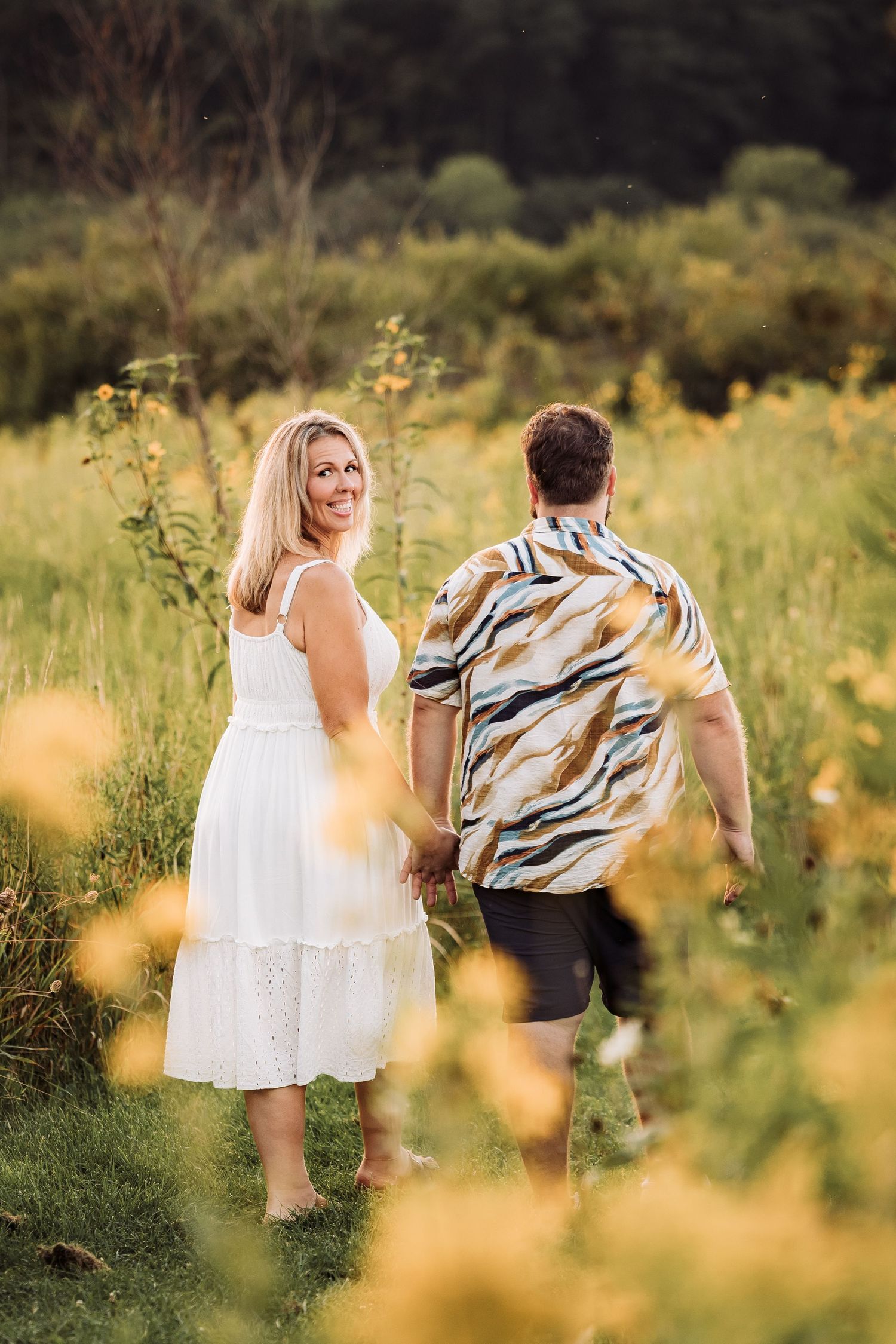 Couple holding hands walking through a sunny wildflower field, woman in white dress looking back and smiling.