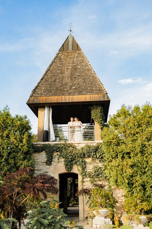 Couple in wedding attire stands on the balcony of a stone tower with a pyramid-shaped wood shingle roof, surrounded by lush green gardens.