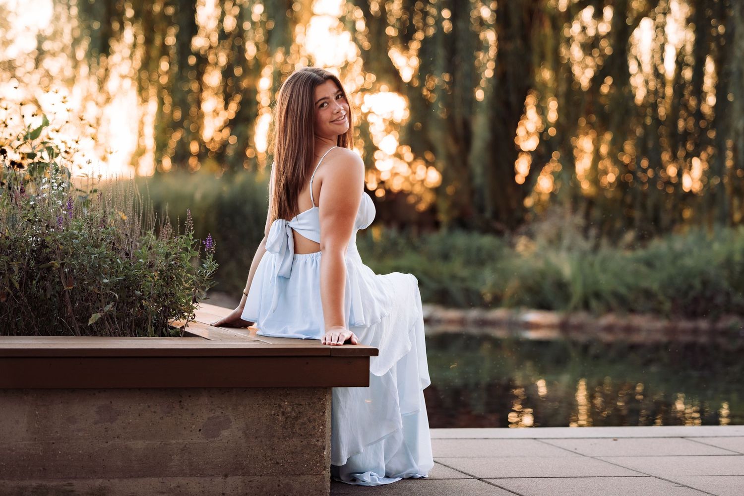 Young woman in white dress sitting on stone bench by pond at golden hour sunset with lush green trees.